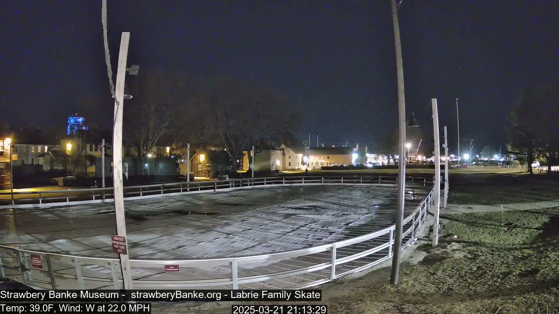 An outdoor ice skating rink at night, under a dark sky with a 22 mph wind, is mostly empty except for some markings on the ice.