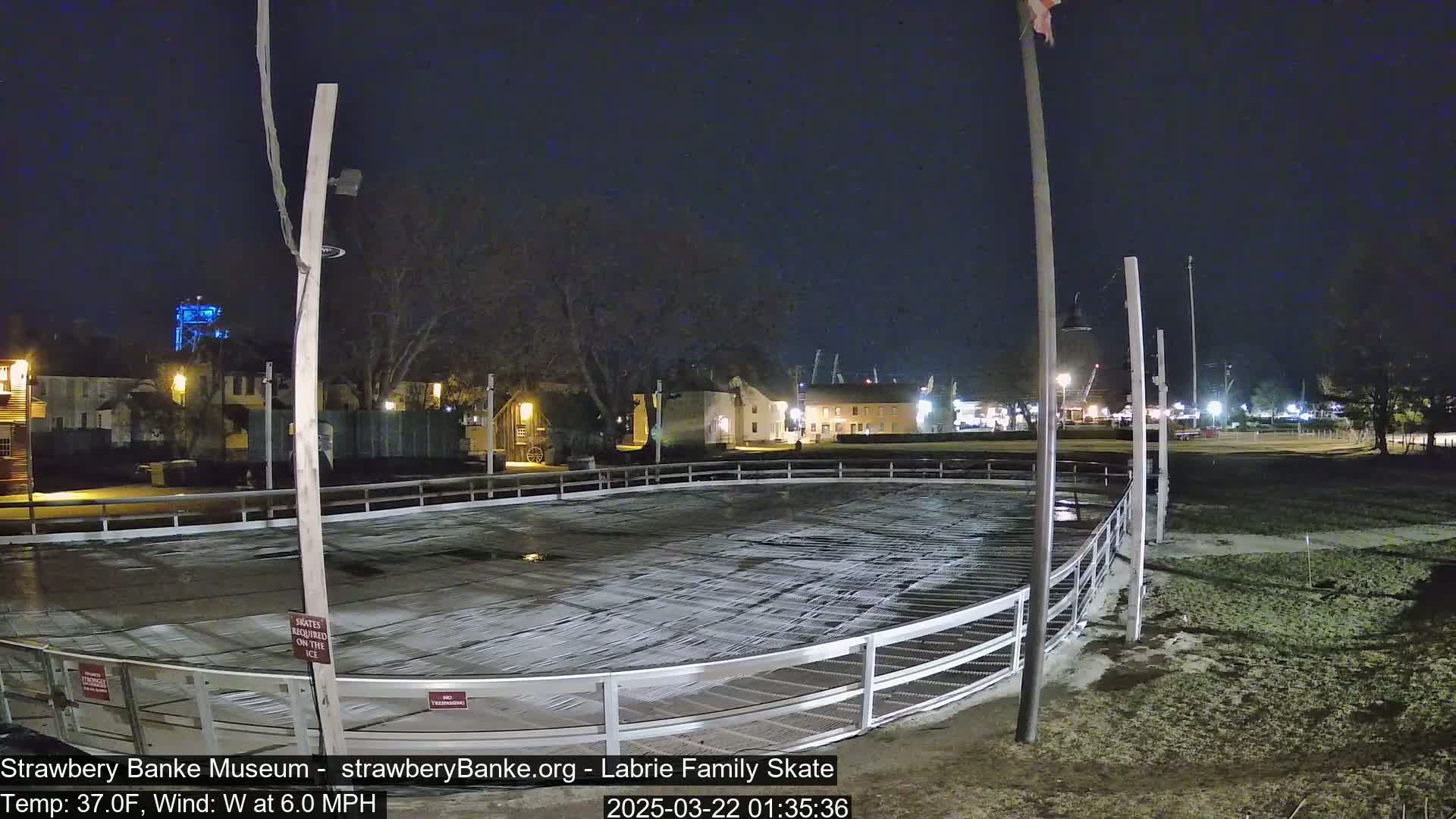 An outdoor ice rink, partially covered in a thin layer of snow, is illuminated at night under a dark sky with a 6.0 mph west wind and a temperature of 37°F.
