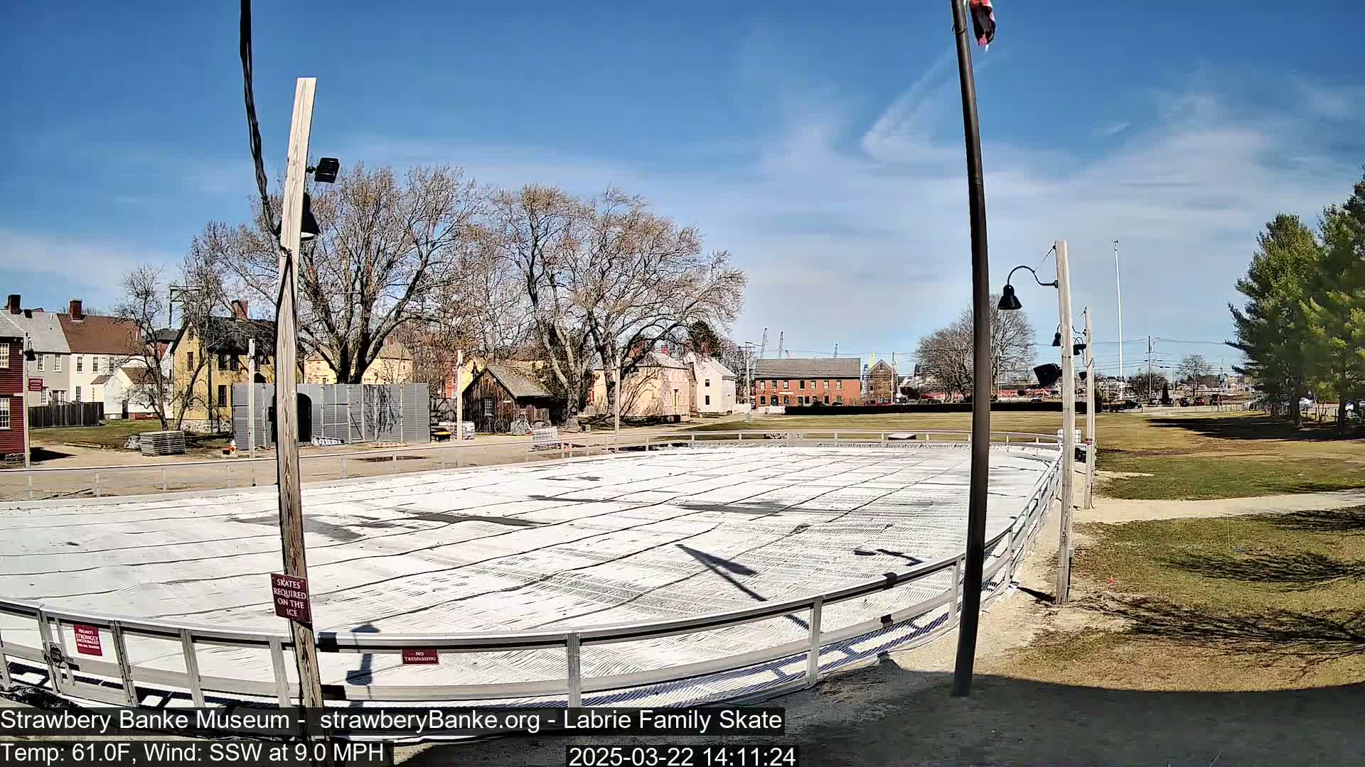 An empty outdoor skating rink, surrounded by bare trees and historical-looking buildings under a sunny sky.