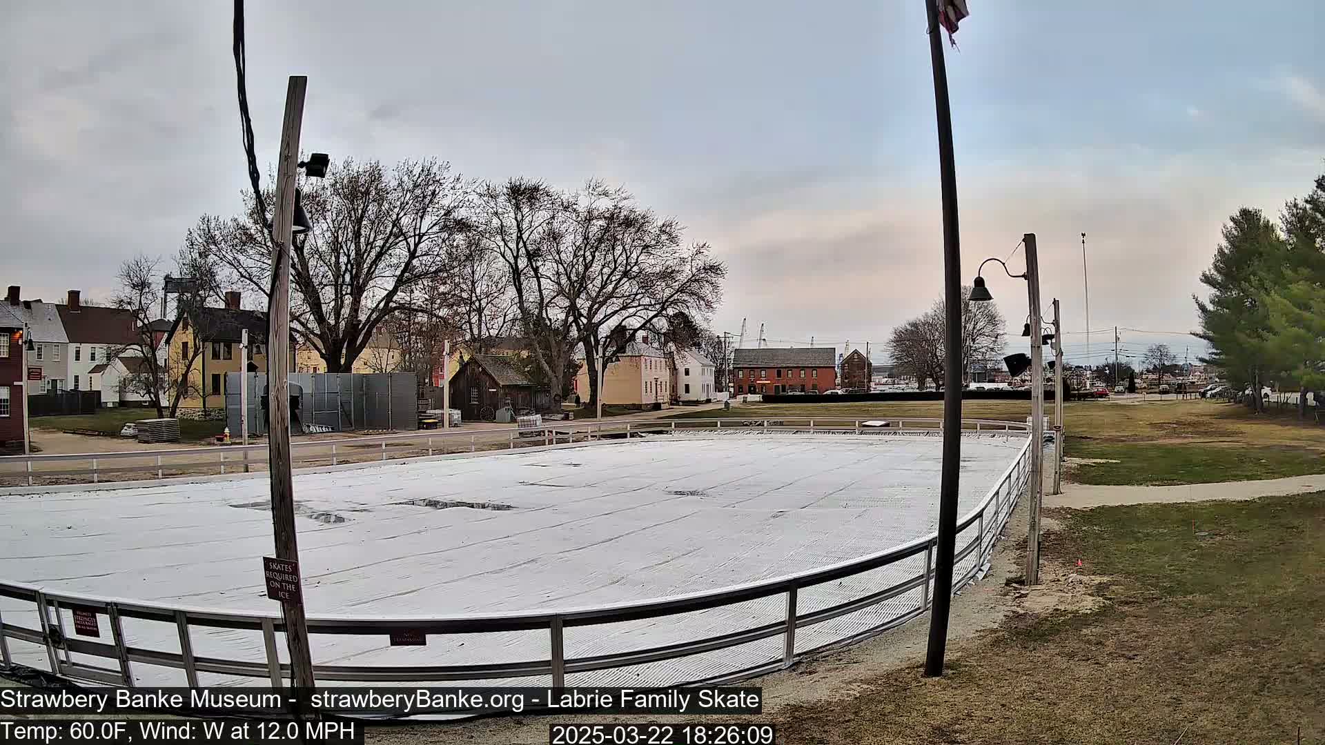 A covered outdoor ice rink sits in a park-like setting next to several buildings under a cloudy sky with a 60°F temperature and a 12mph west wind.