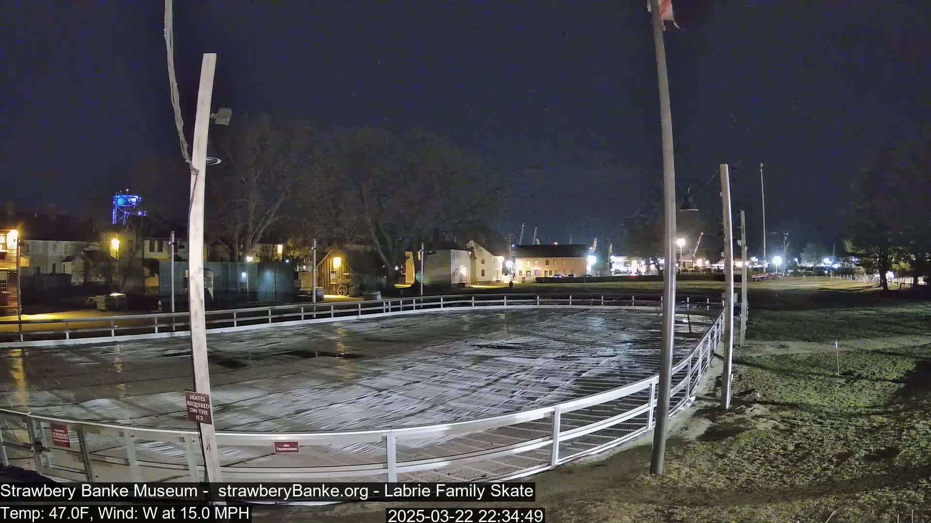 An empty outdoor ice skating rink at night under a dark sky with a 15 mph wind.