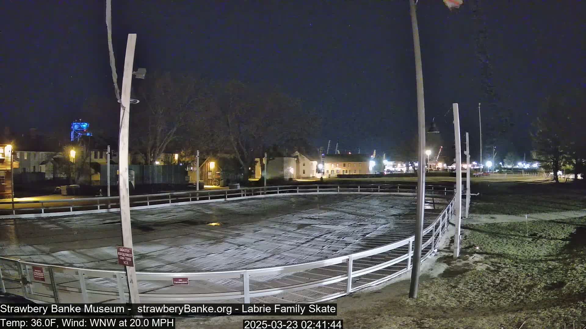 An outdoor ice rink is mostly empty at night under a clear, dark sky, with a 20 mph wind, and buildings and lights visible in the background.