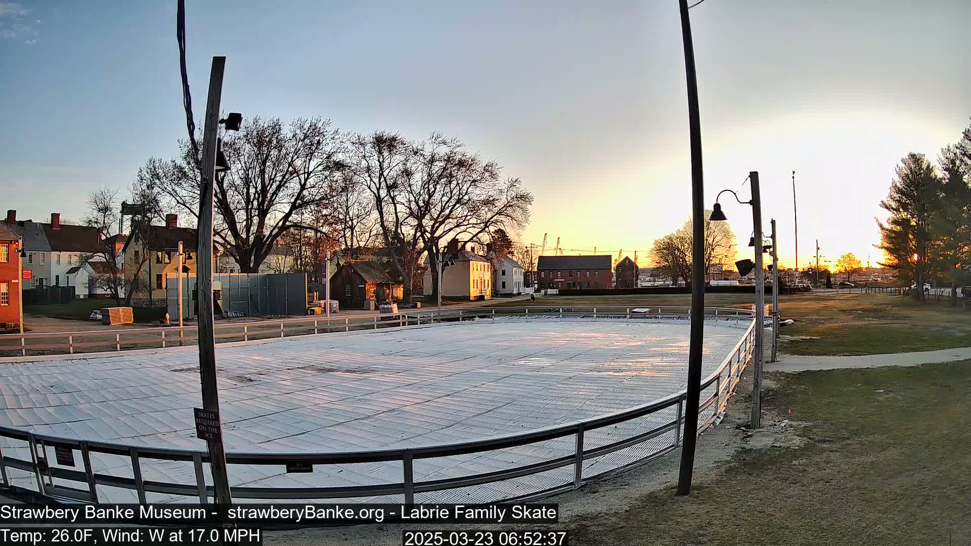 An outdoor ice rink sits near several historical-looking buildings at sunset, under a clear sky with a 17 mph wind and a temperature of 26 degrees Fahrenheit.