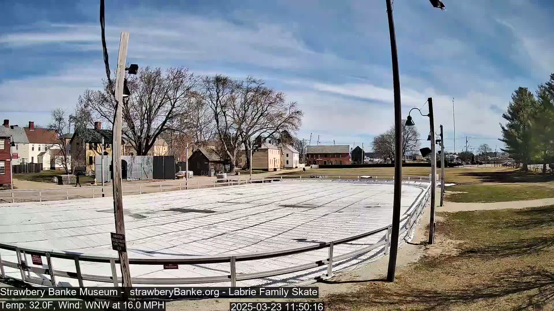 A covered outdoor ice rink sits in a grassy area near several historic-looking buildings under a mostly sunny sky with a 16 mph wind.
