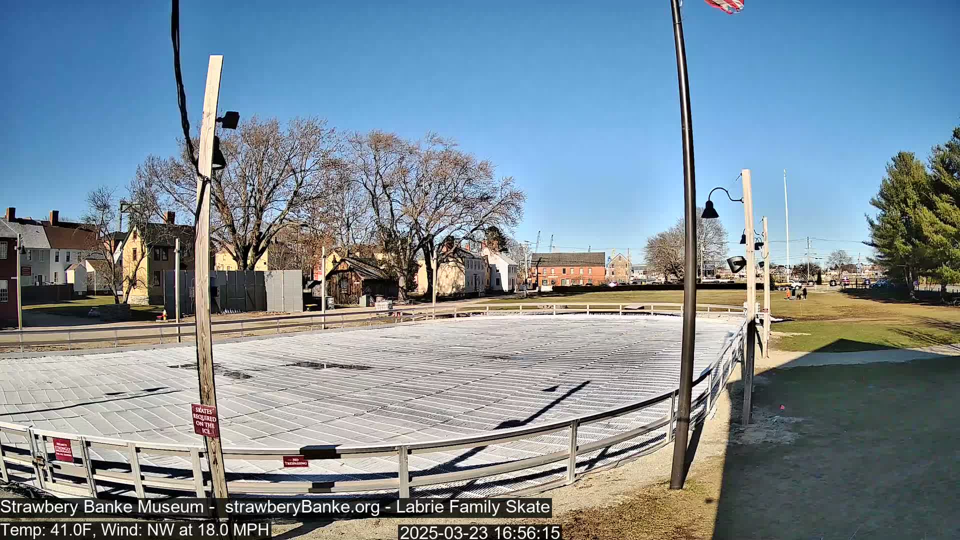 An outdoor ice skating rink, surrounded by a metal fence and situated near historical-looking buildings under a sunny sky.