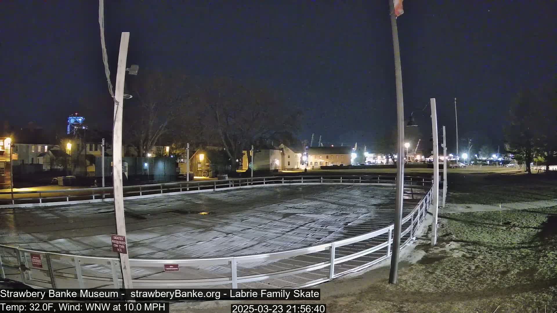 An outdoor ice rink, mostly empty, is illuminated at night under a dark sky with a wind of 10 mph.