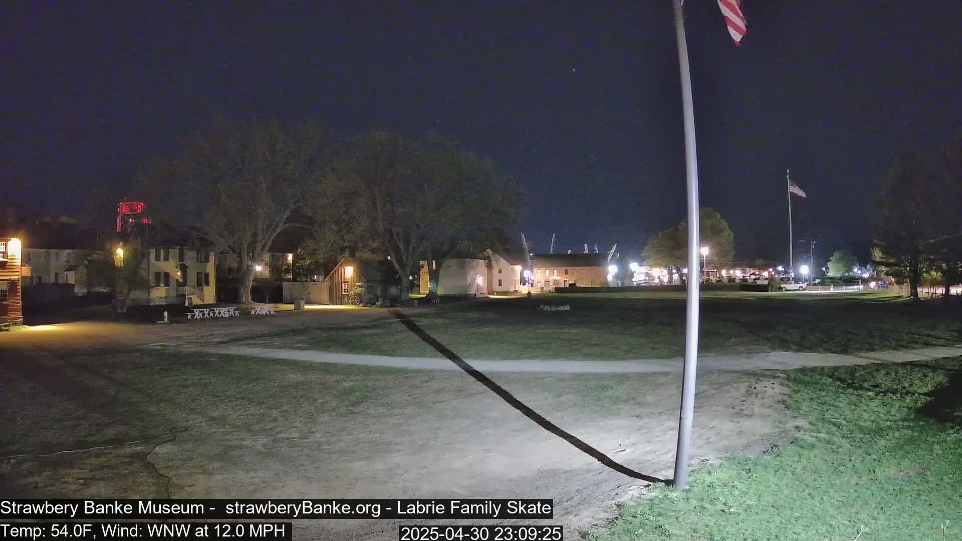 A nighttime view of a grassy area with historical-looking buildings and a flagpole, under a clear, dark sky with a 12 mph wind.