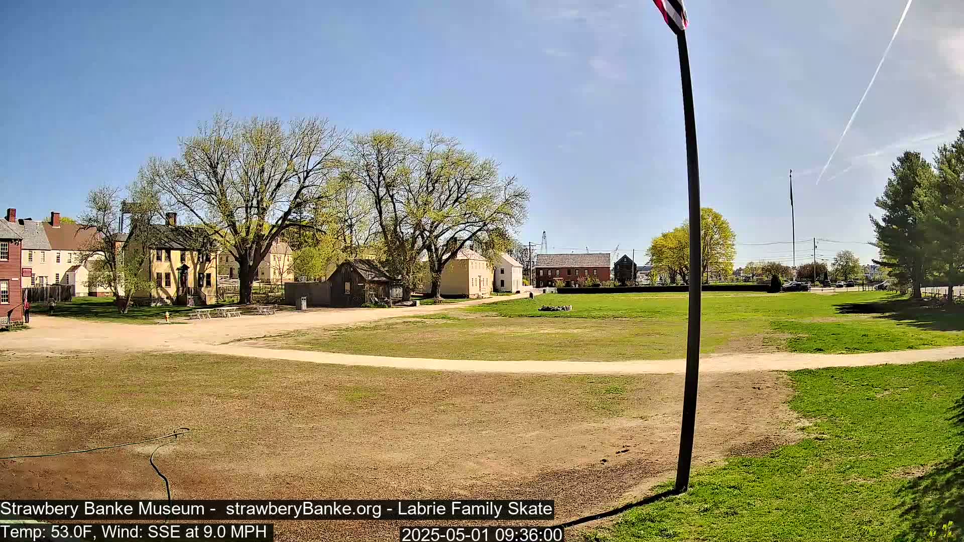 A sunny, breezy day reveals a grassy field and several historical-looking buildings in a colonial-style setting.