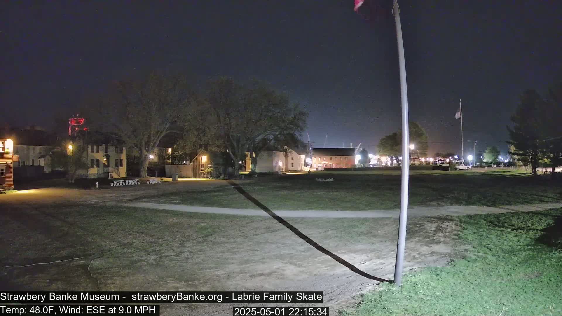 A nighttime view of a grassy area with several historical-looking buildings and a tall flagpole, under a dark sky with a wind of 9.0 mph from the east-southeast.