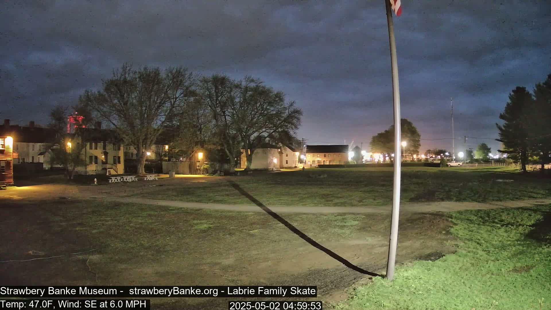 A nighttime view of a grassy area with historical-looking buildings and trees under a dark, cloudy sky.