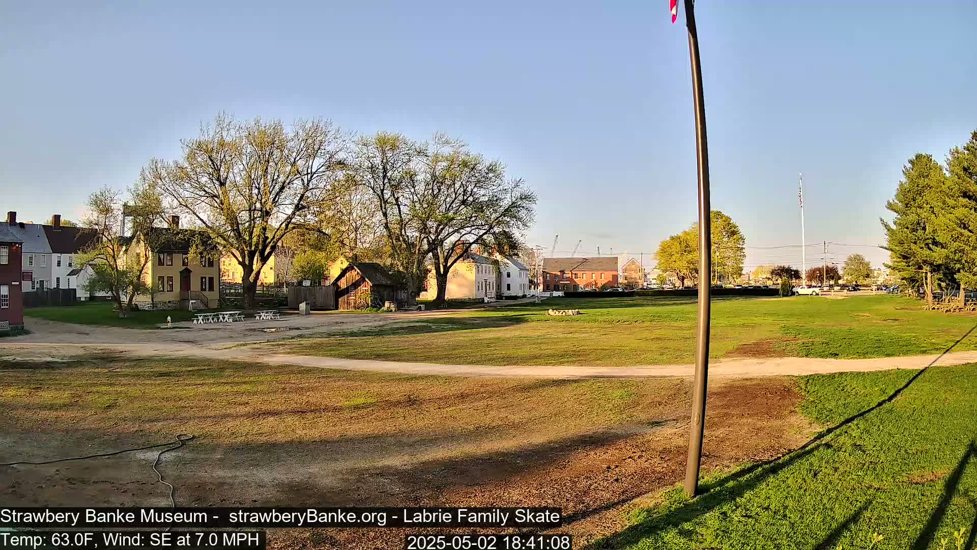 A grassy field with several historical-looking buildings and trees in the background, under a clear, sunny sky with a slight southeast wind.