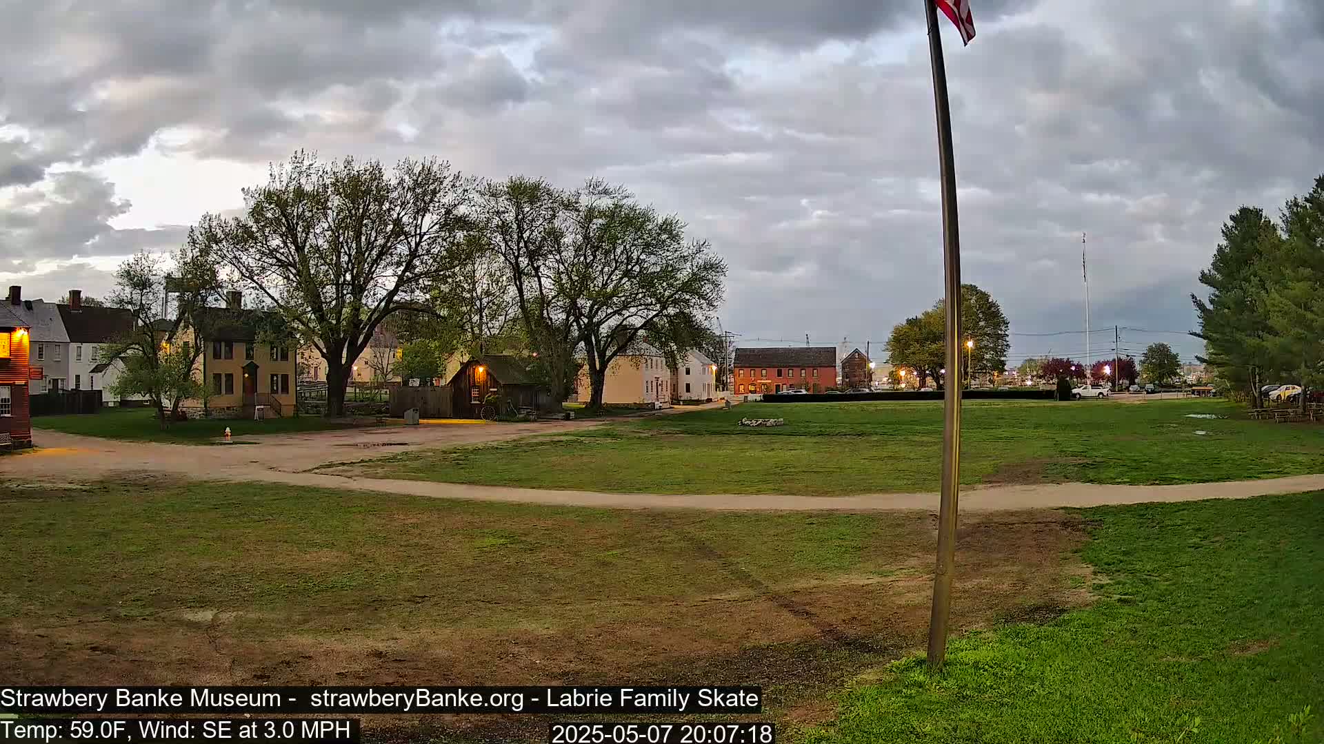 A grassy field with historical-looking buildings and trees in the background under a cloudy evening sky.