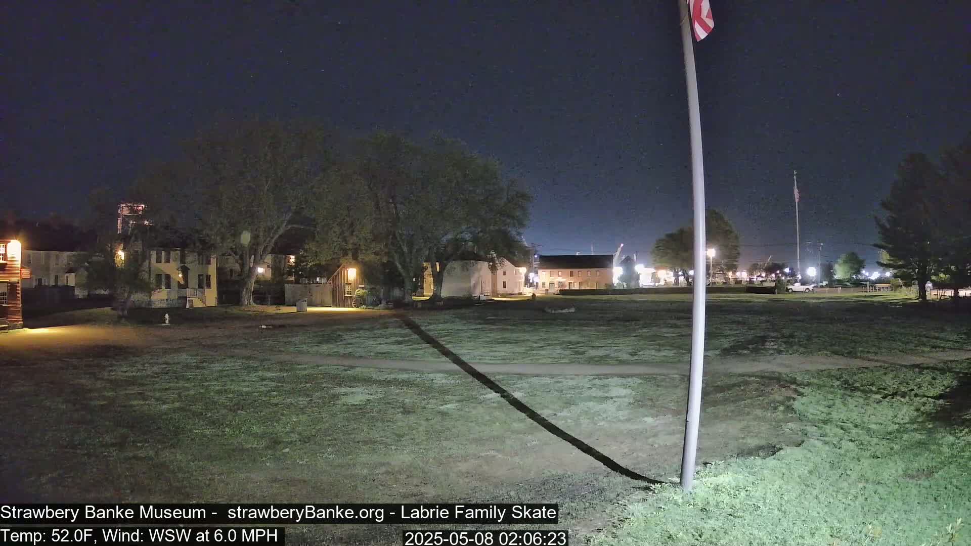 A nighttime view of a grassy area with historical-looking buildings in the background under a clear, dark sky.