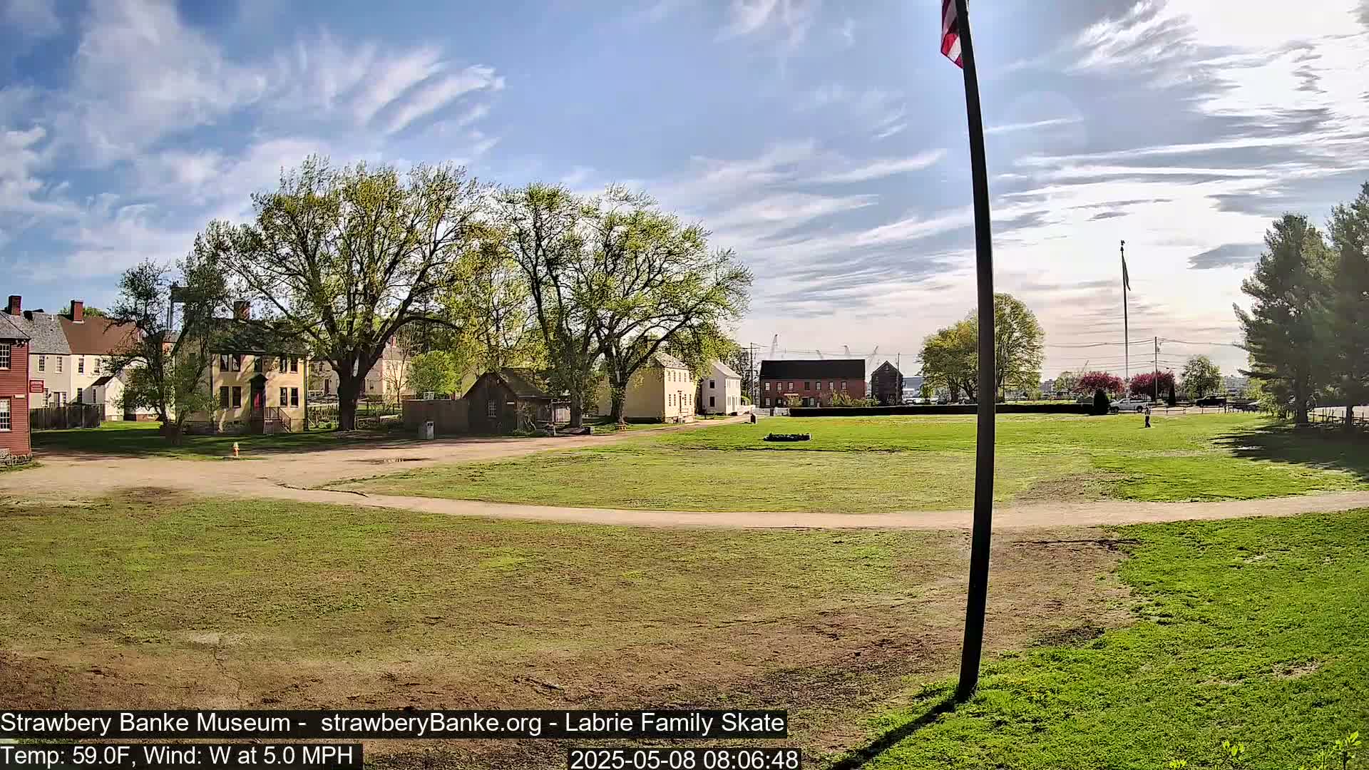 Under a partly cloudy sky, a grassy field stretches out before several historic-looking buildings, with a flagpole standing tall in the foreground.