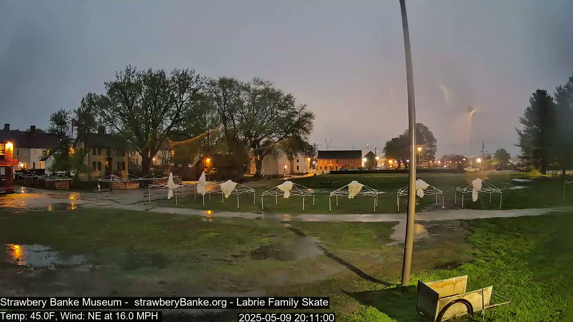 A wet, grassy area with several overturned canopies and historical-looking buildings in the background under a stormy, nighttime sky with a visible lightning strike.