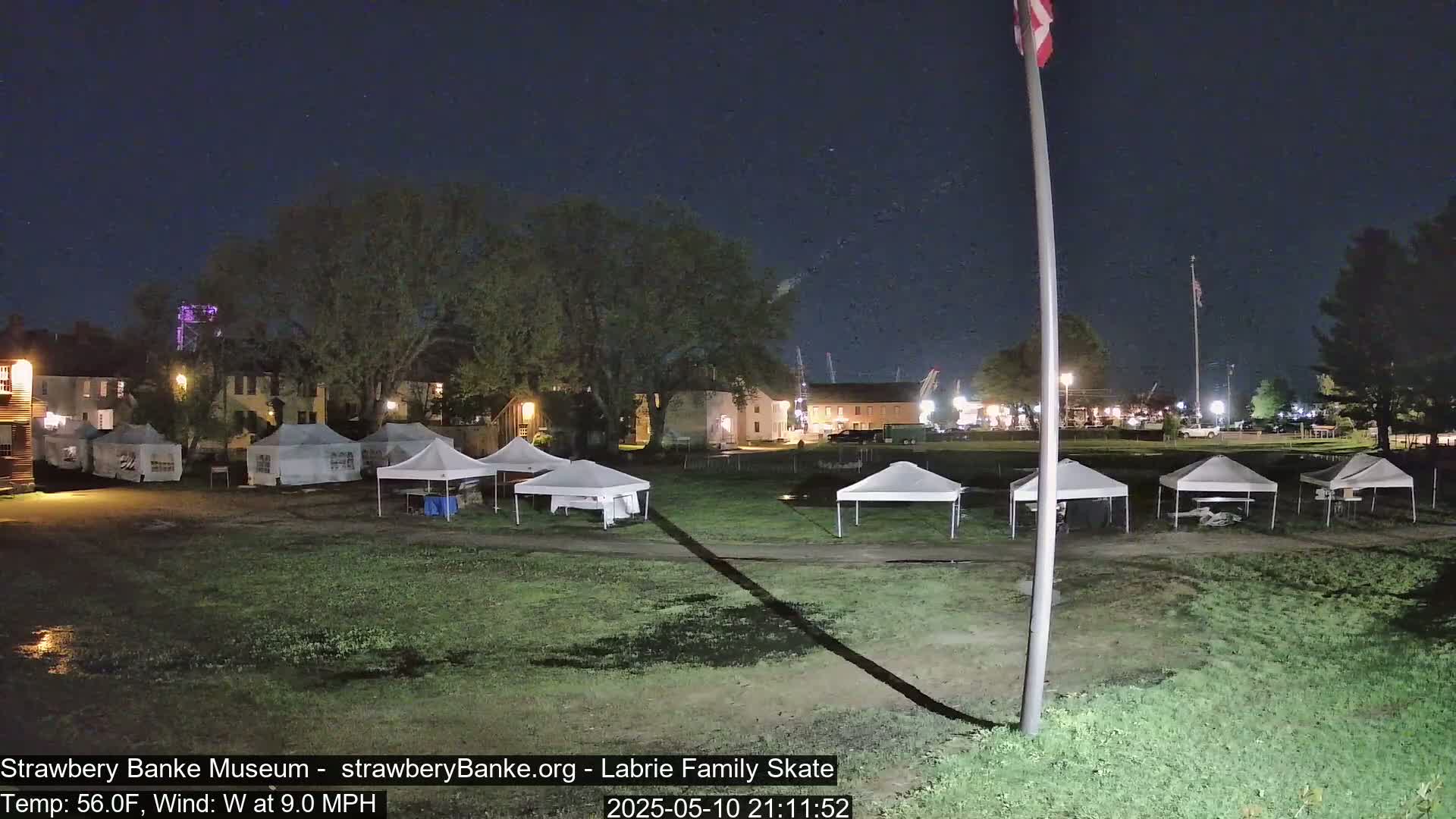 At night, under a dark sky with a west wind, several white canopies are set up on a grassy area in front of historic-looking buildings.