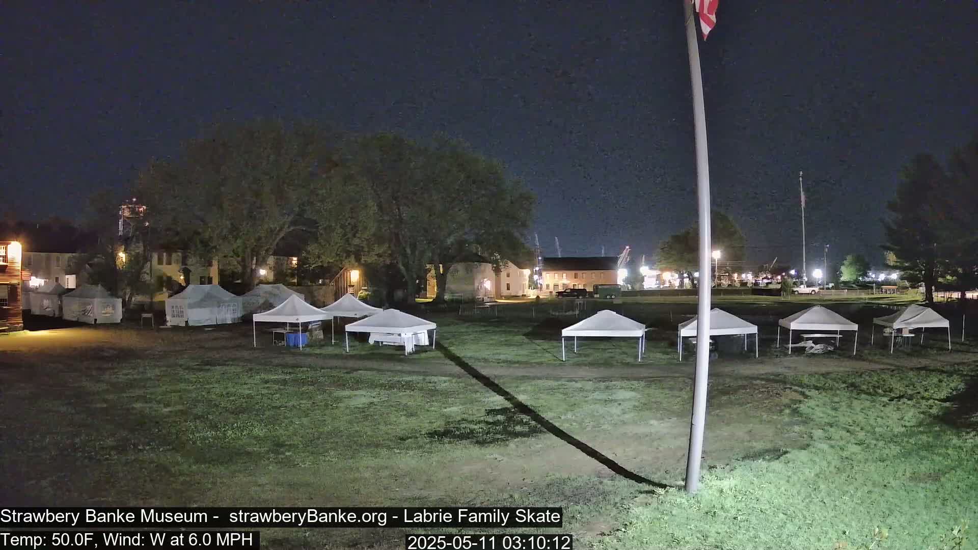A nighttime outdoor scene shows several white canopy tents set up on a grassy area in front of buildings, under a clear, dark sky with a light west wind.