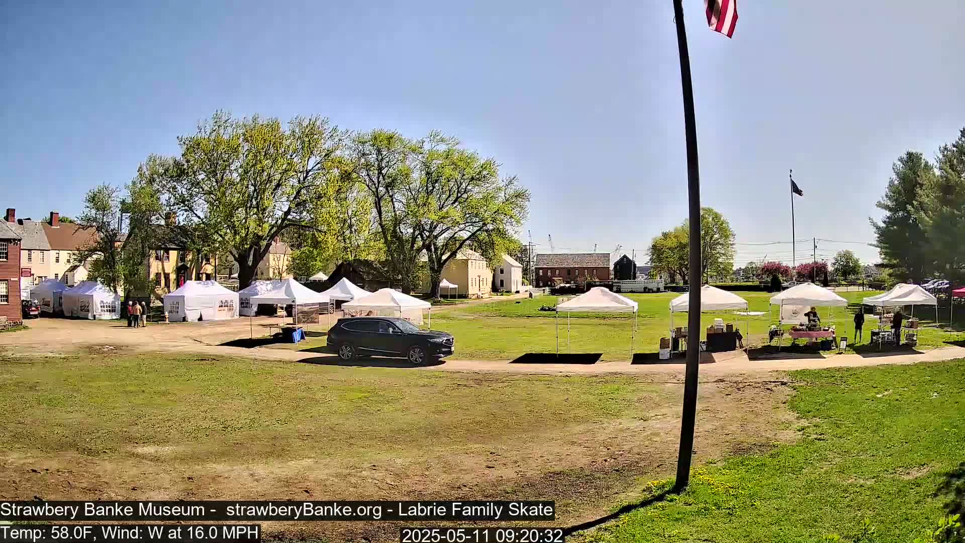 A sunny day with a west wind at 16 mph shows several white canopies set up in a grassy area near historic buildings, with a dark-colored SUV parked nearby.