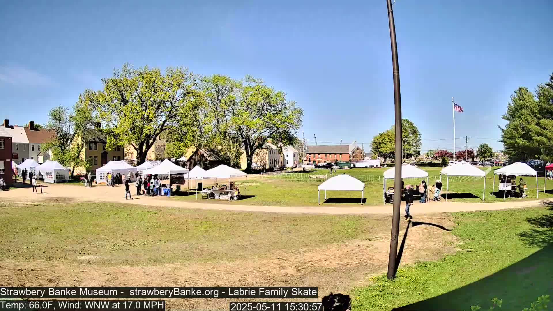 A sunny outdoor market scene with numerous white canopy tents set up on a grassy field, surrounded by historic buildings and trees, under a clear blue sky.