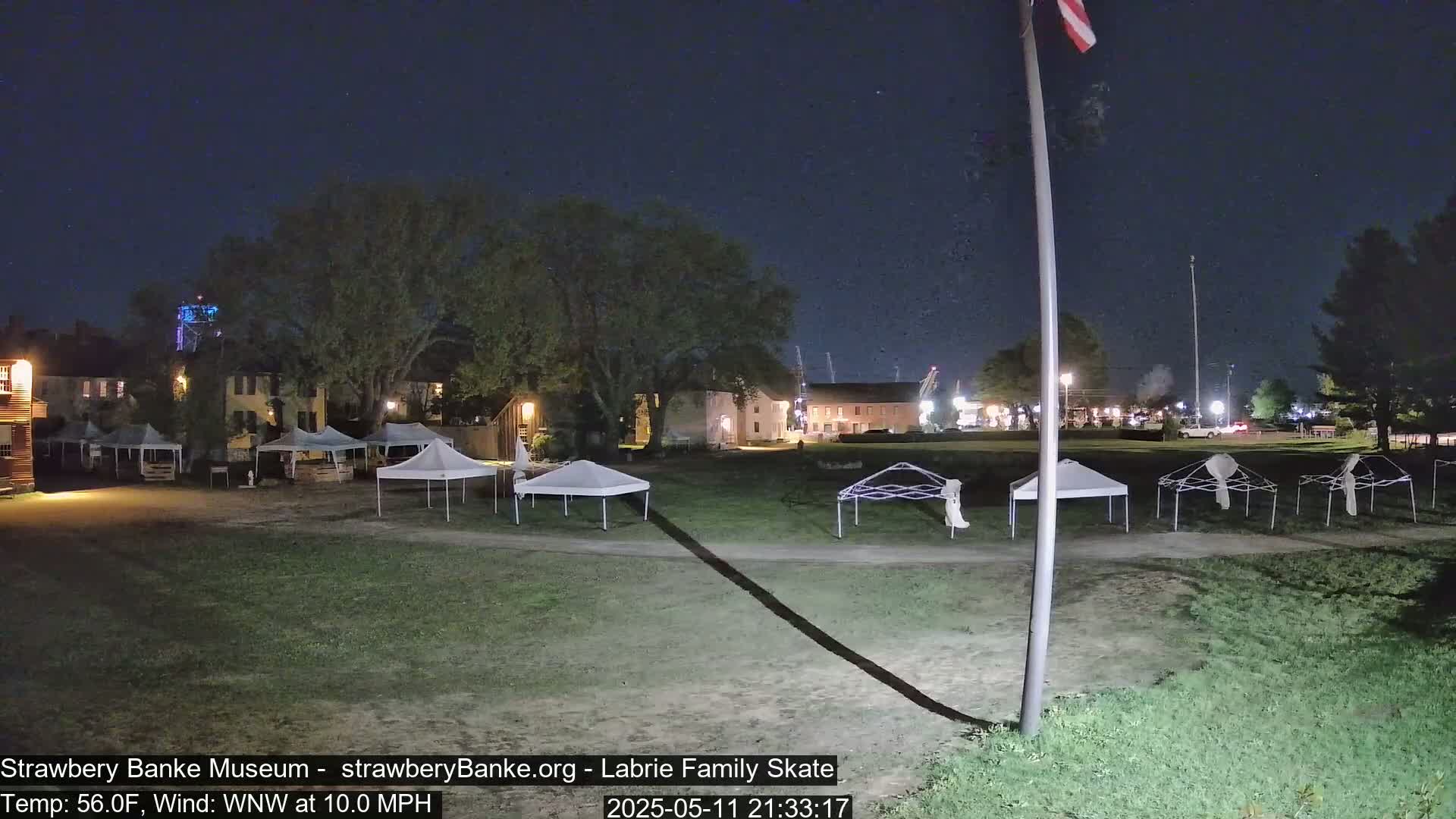 At night, under a clear sky with a 10 mph wind, several white canopy tents are set up on a grassy area in front of historic-looking buildings.