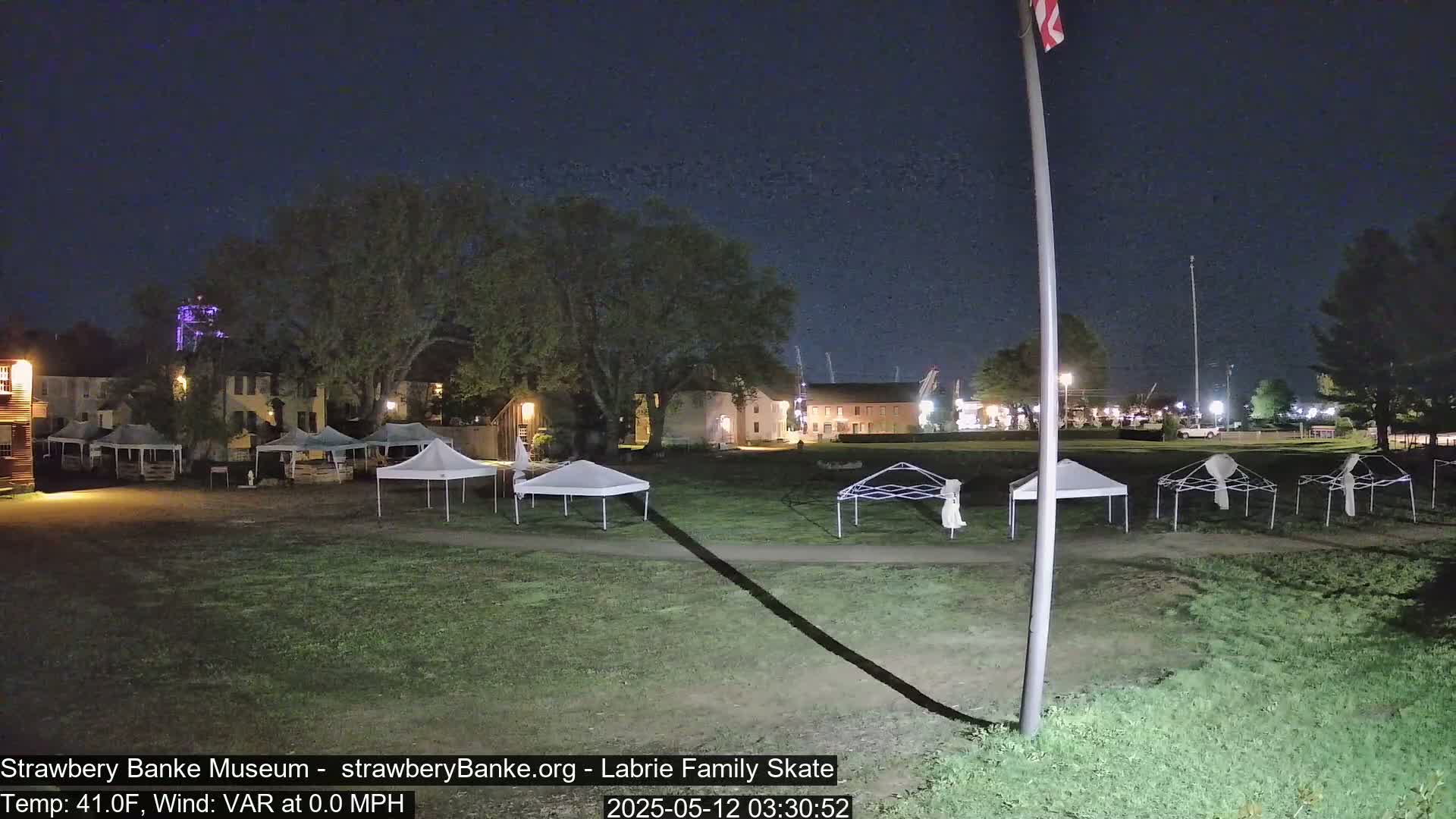 At night, several white canopies are set up on a grassy area in front of historical-looking buildings under a clear, dark sky.