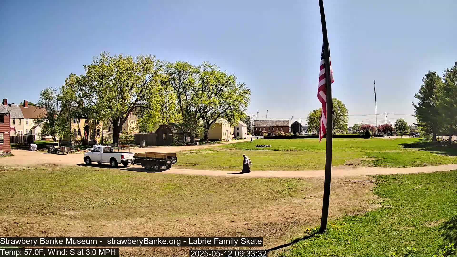 A person walks on a dirt path across a grassy field, near several historical-looking buildings and a pickup truck towing a flatbed trailer, under a sunny sky.