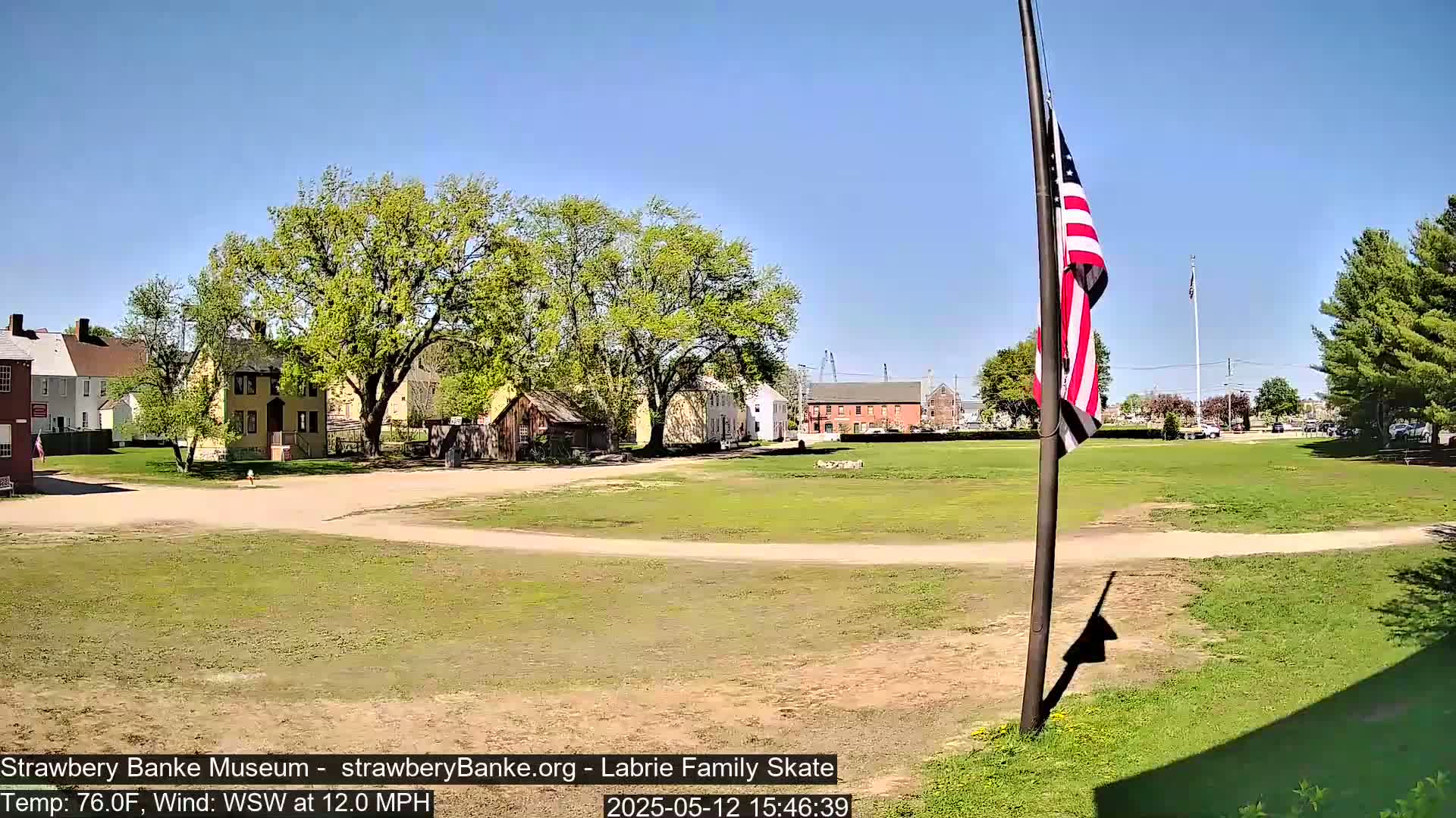 A grassy field with historical-looking buildings and a large American flag on a pole, under a clear blue sky on a sunny day with a 12 mph wind.