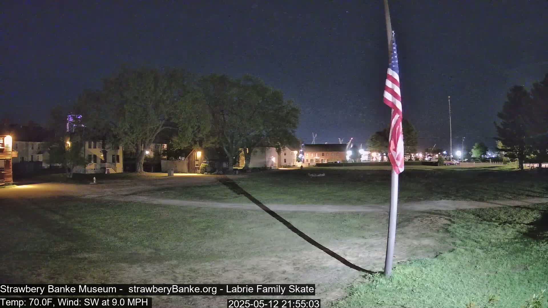 A nighttime view of a grassy area with several historic-looking buildings in the background, under a clear sky, with a flagpole and American flag in the foreground, and a southwest wind.