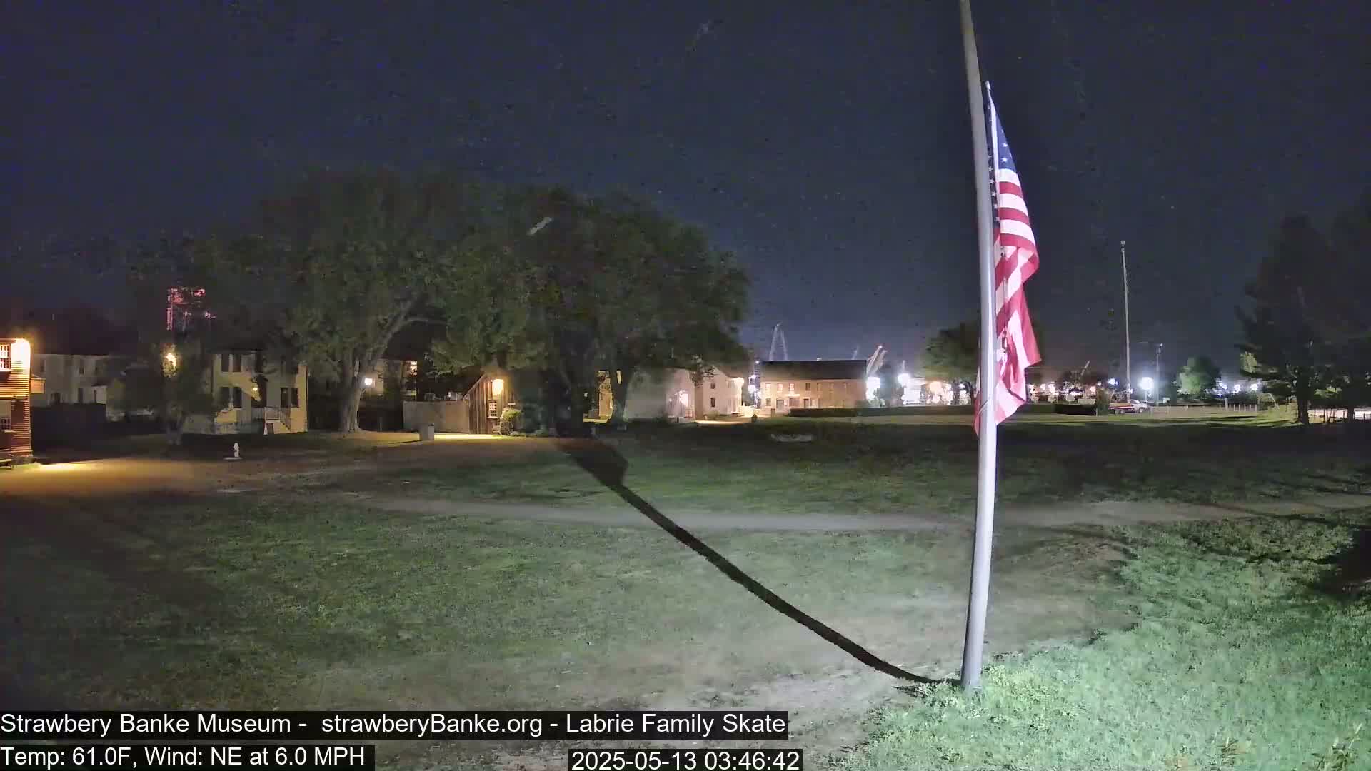 At night, under a dark sky with a northeast wind, a grassy area with several historical-looking buildings is illuminated, and an American flag stands tall on a pole in the foreground.