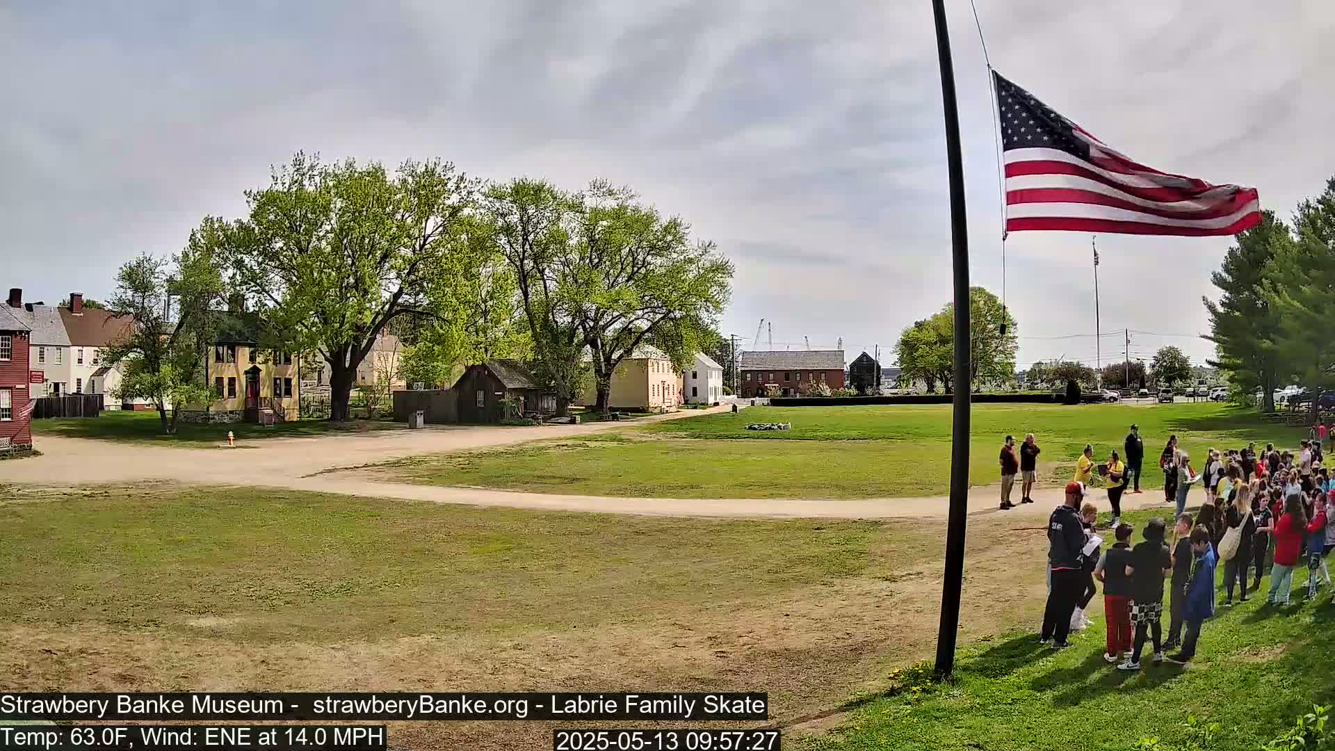 A large group of people stands on a grassy field in front of several historical-looking buildings under a partly cloudy sky with a flagpole displaying an American flag, a light breeze blowing.