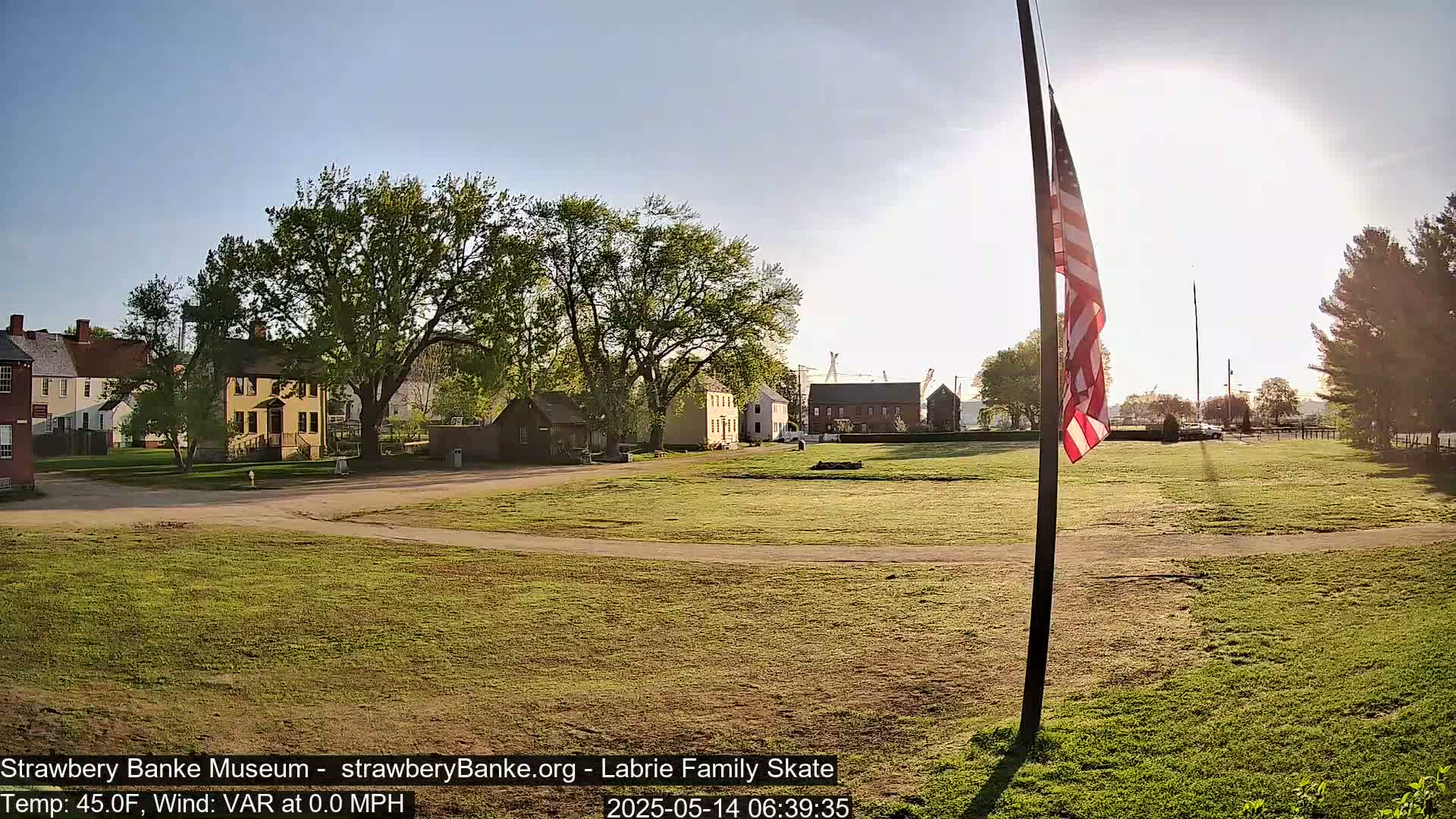 A grassy field with historical buildings and a US flag on a pole in the sunny morning.