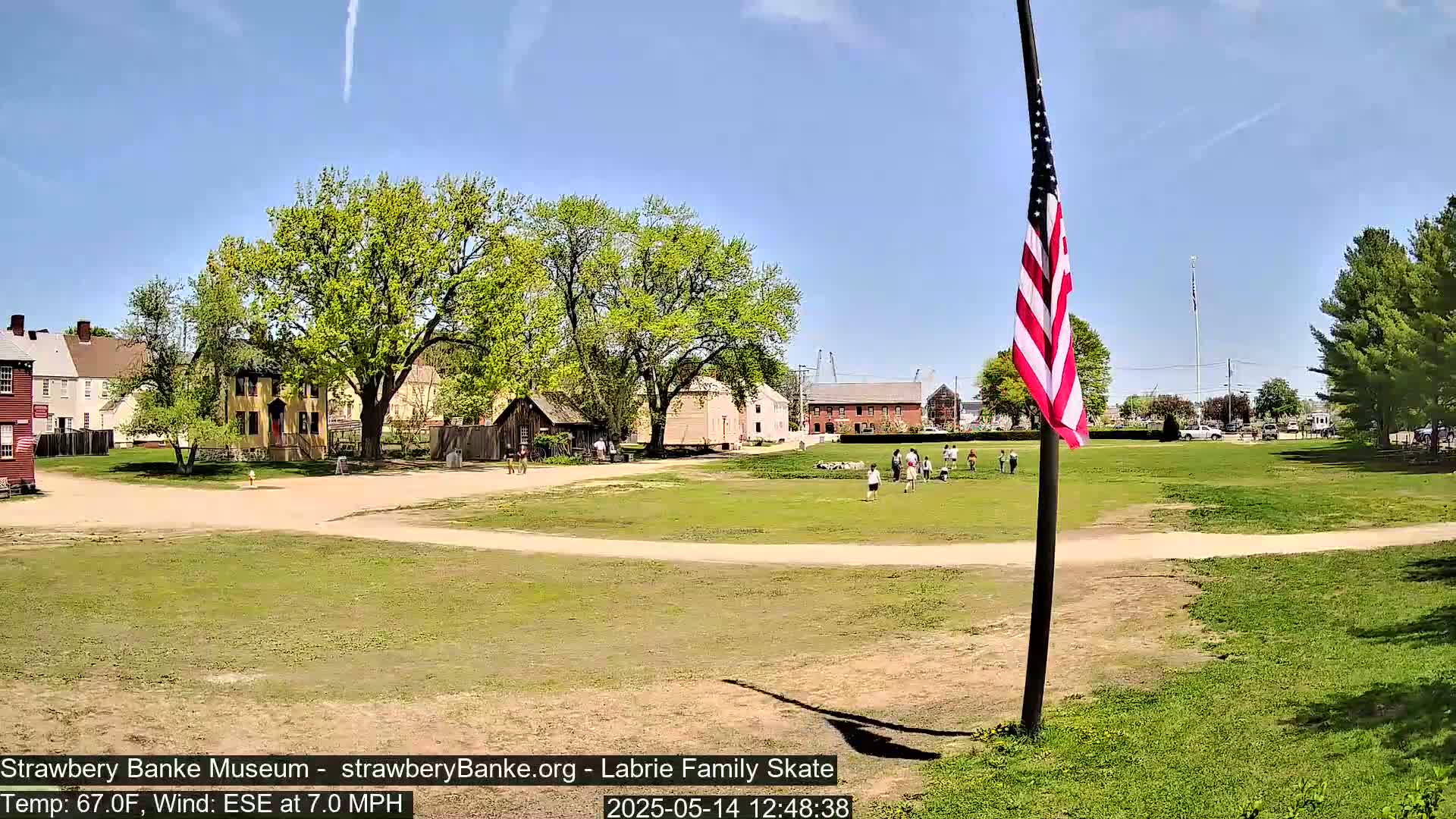 A grassy field with several historical-looking buildings and a few people, under a sunny sky with a light wind.