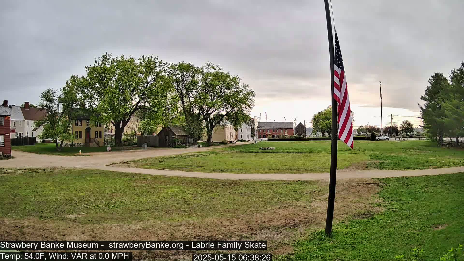 A grassy field with several historical-looking buildings and a large American flag on a pole under an overcast sky.
