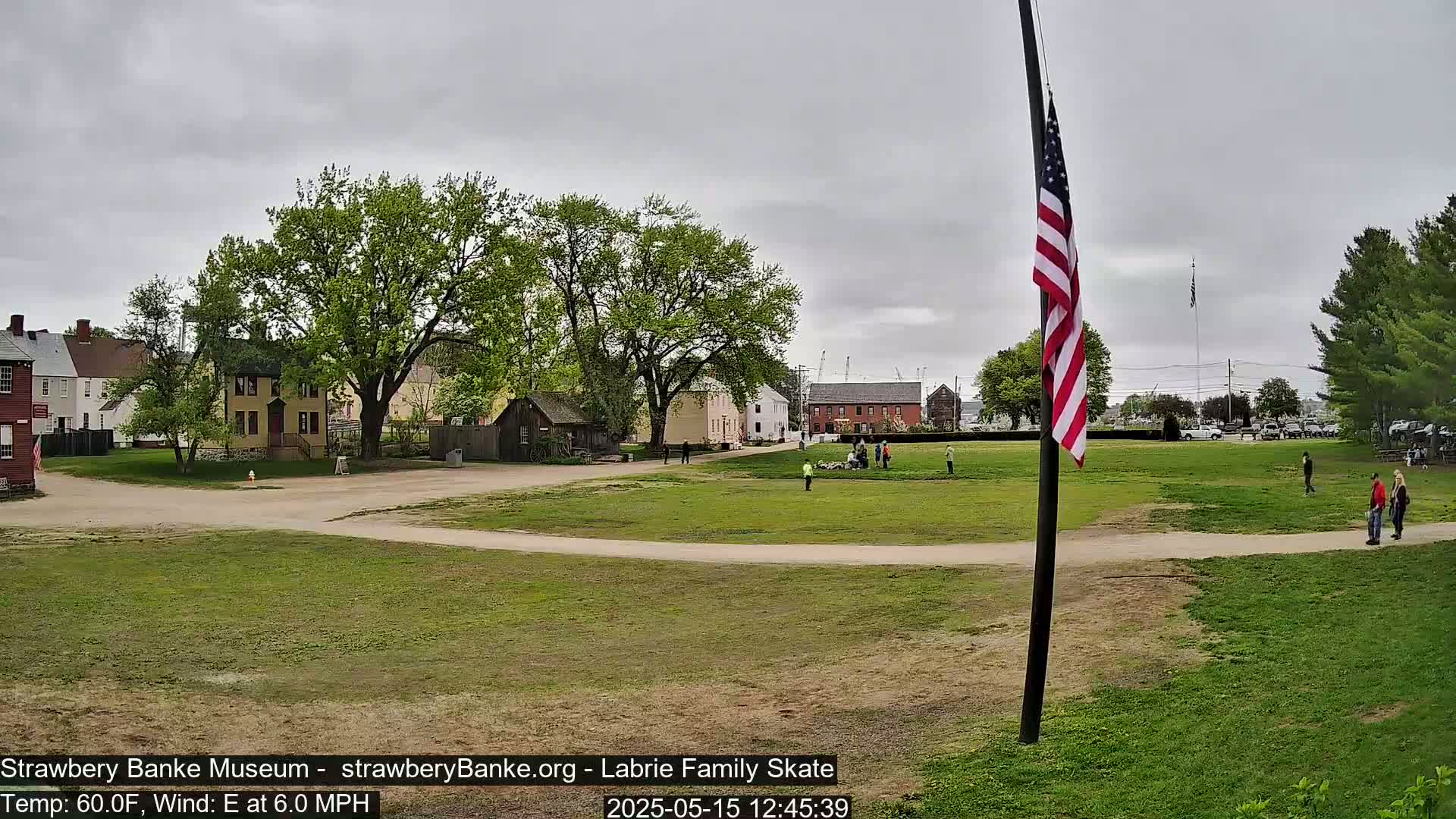 A grassy field with several historical-looking buildings and a few people, under an overcast sky.