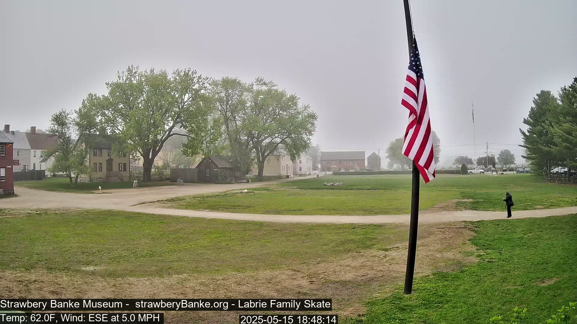 A person stands in a grassy field next to a dirt path, in front of several historical-looking buildings on a foggy day.