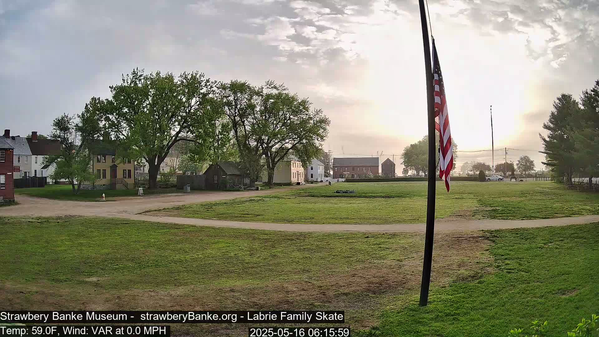 A grassy field with historical-looking buildings and a waving American flag under a mostly cloudy, sunrise sky.