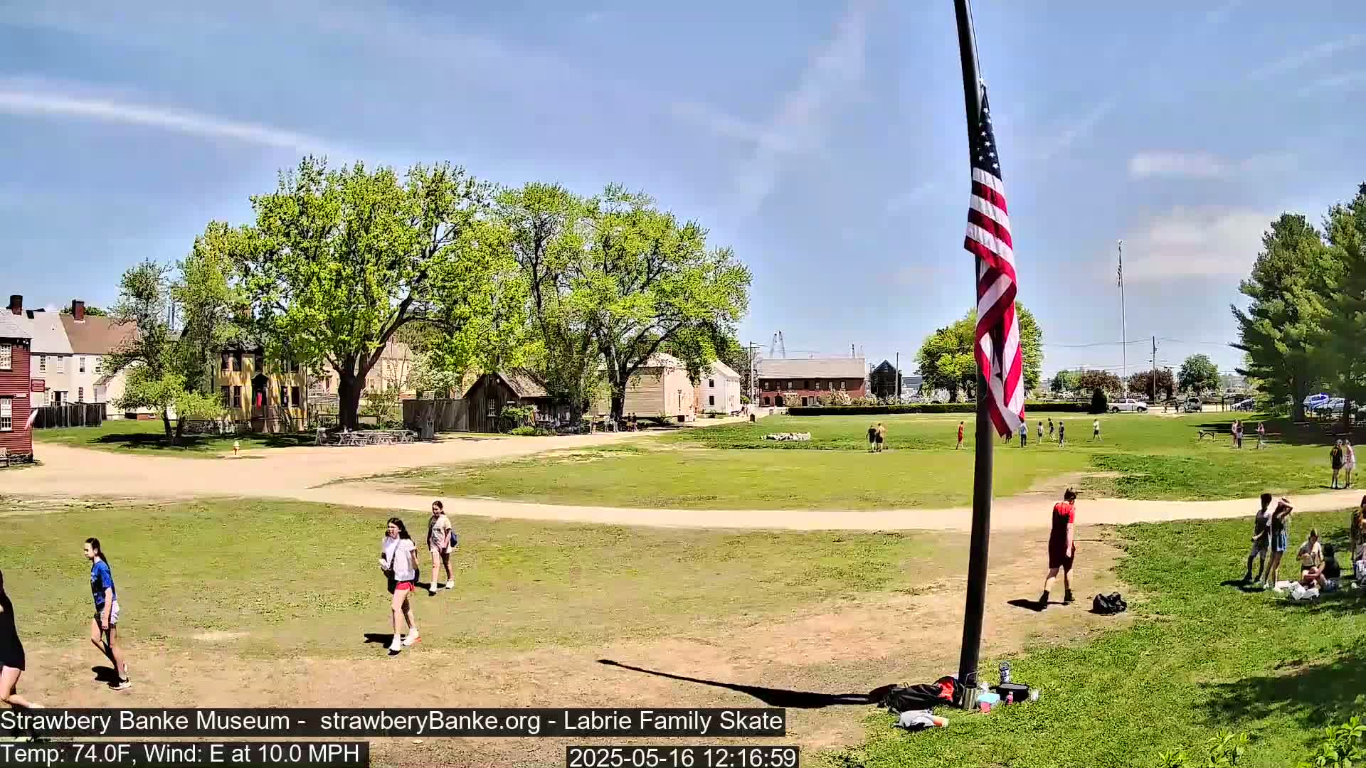 A sunny day with an east wind at 10 mph shows a grassy field with several people walking and sitting near historical-looking buildings and a tall flagpole with an American flag.