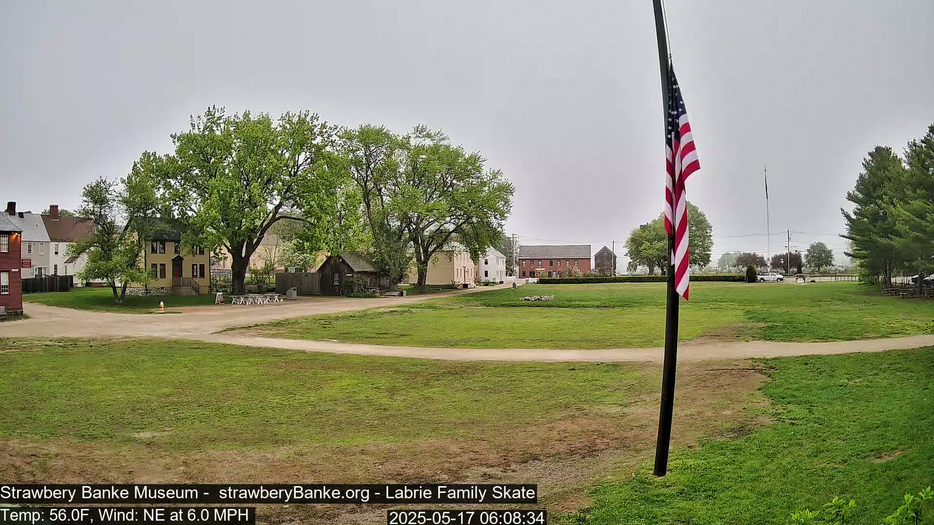 A grassy field with historical-looking buildings and a flagpole displaying an American flag under an overcast sky.
