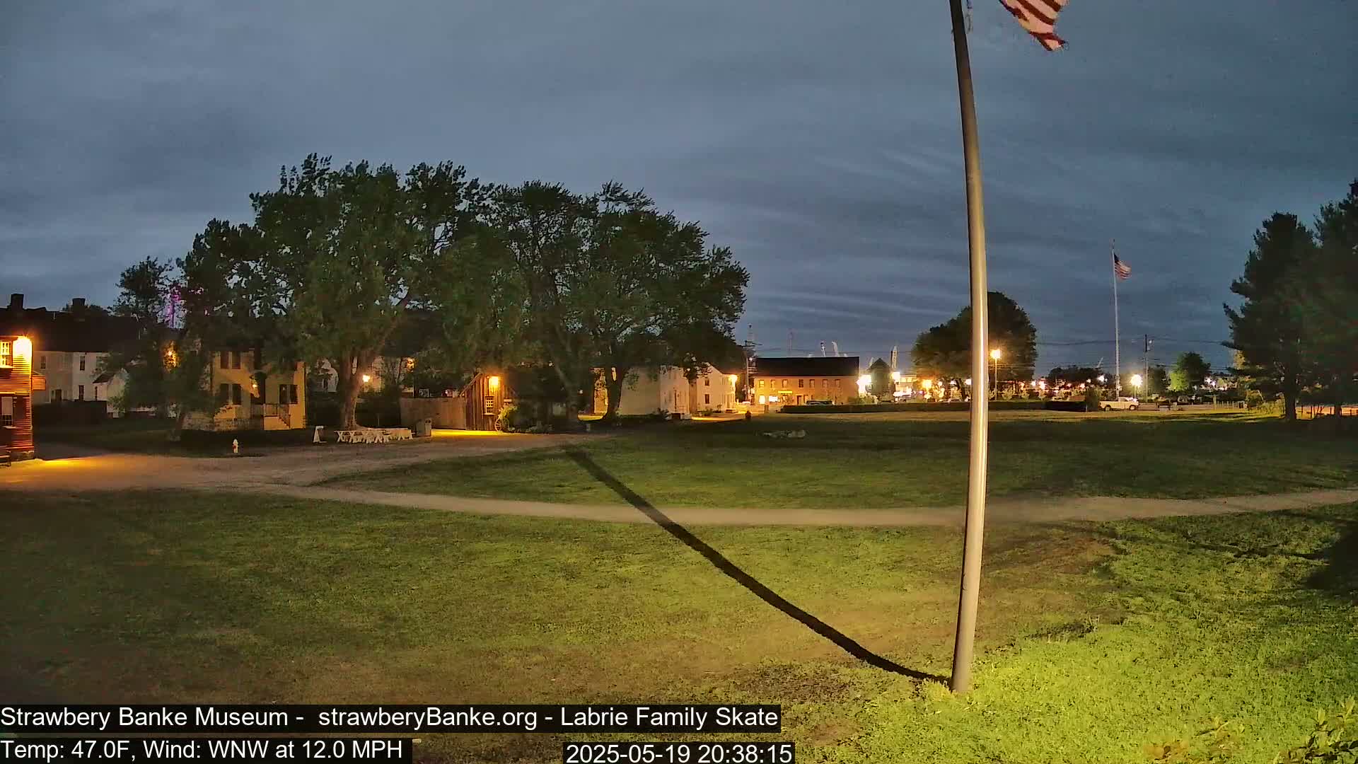 A nighttime view of a grassy field with historical-looking buildings and a flagpole in the background, under a mostly clear twilight sky.