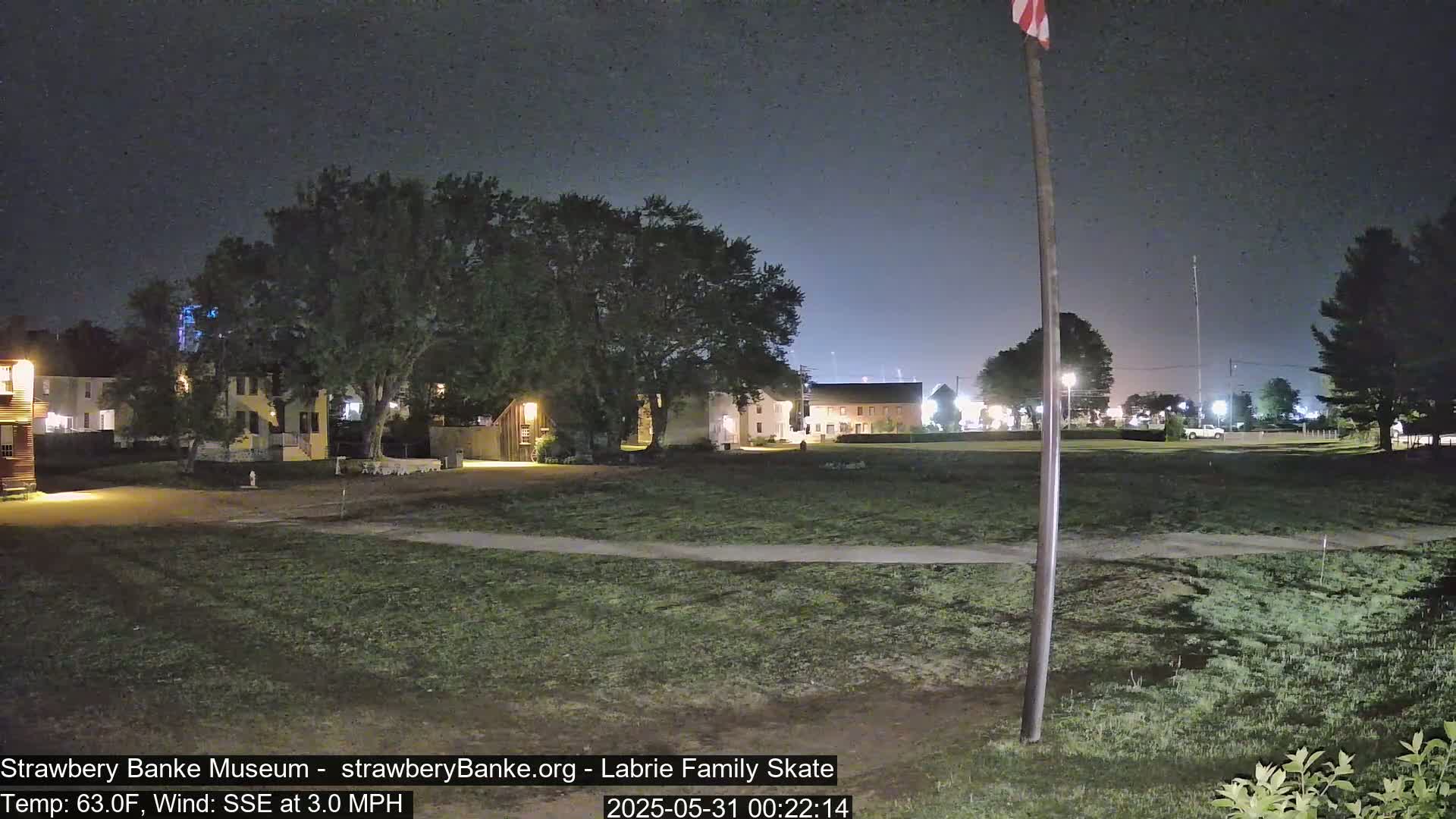 At night, under a dark sky with a slight wind, several illuminated buildings are visible across a grassy area.