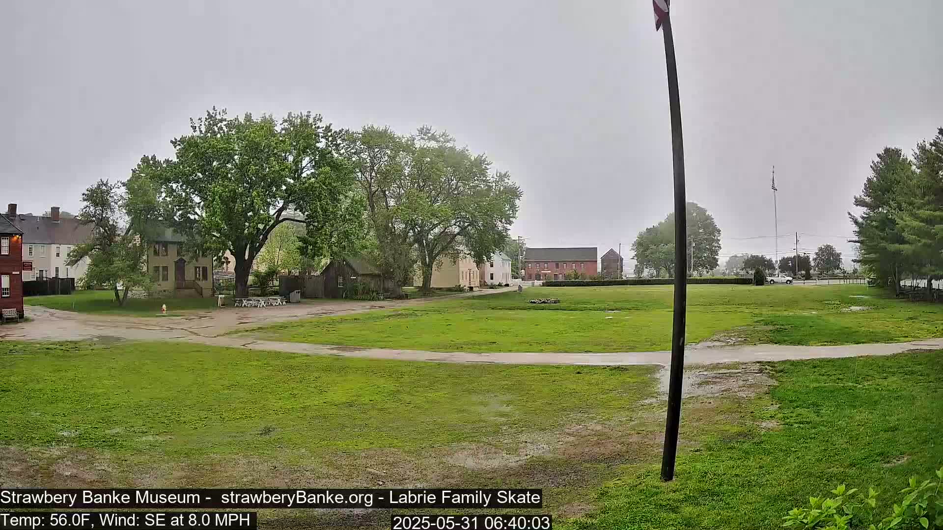 A grassy area with several historical-looking buildings and trees is shown under an overcast sky with light rain.