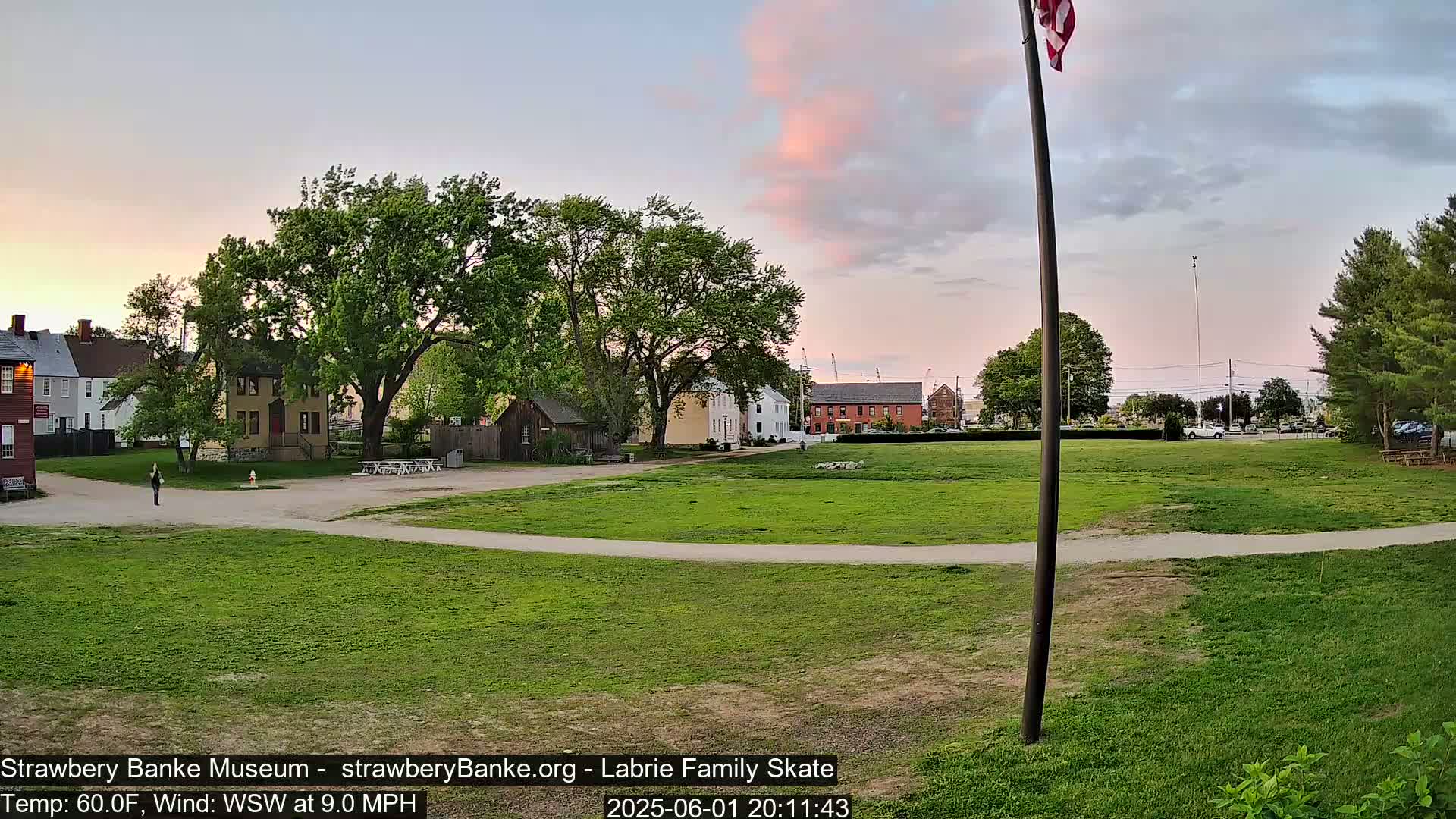 A grassy field with a paved path, surrounded by historical-looking buildings and lush green trees under a partly cloudy, pinkish sunset sky.