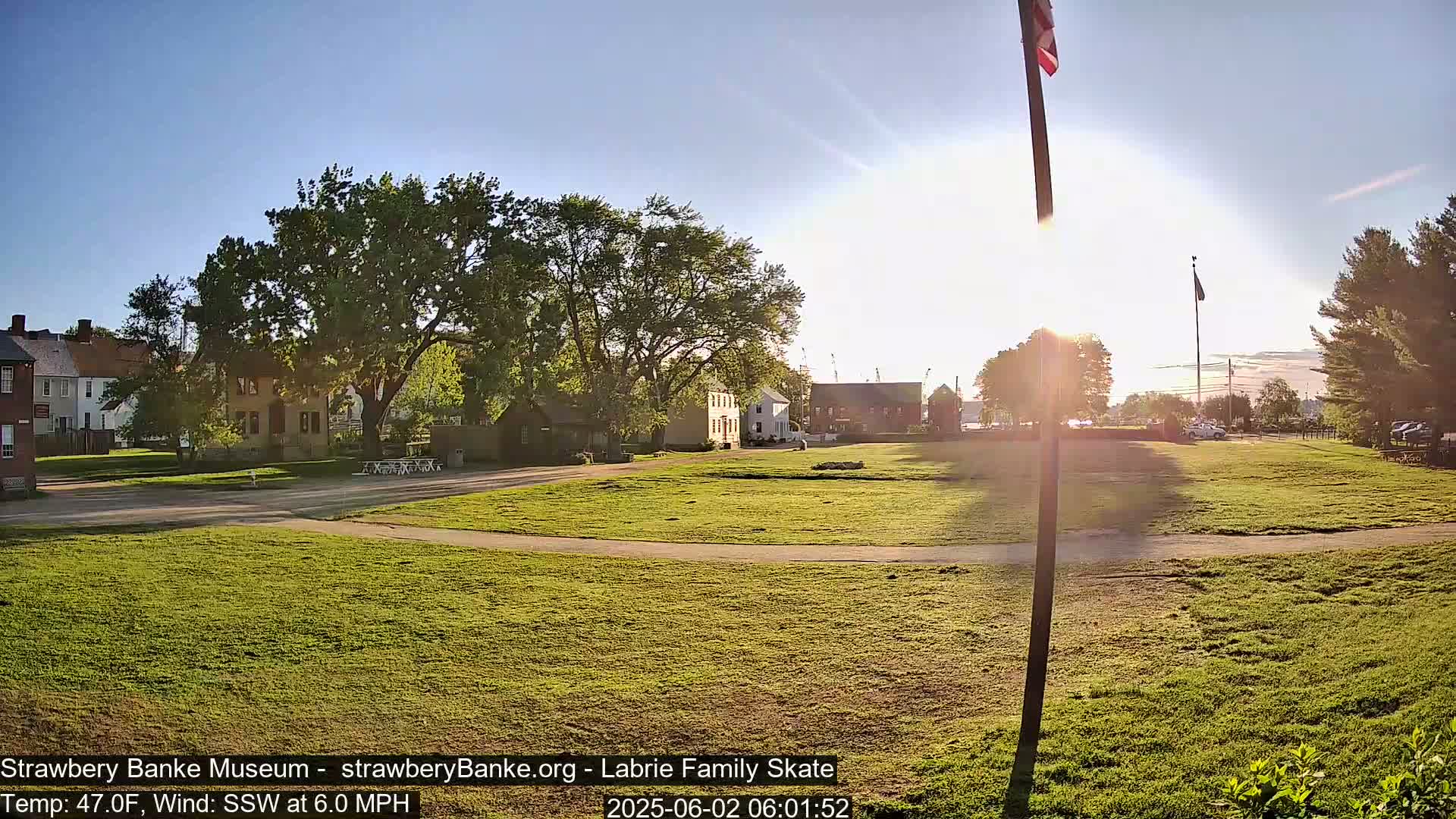 A grassy field with several historical-looking buildings in the background is bathed in the morning sunlight under a clear sky, with a light south-southwesterly wind.