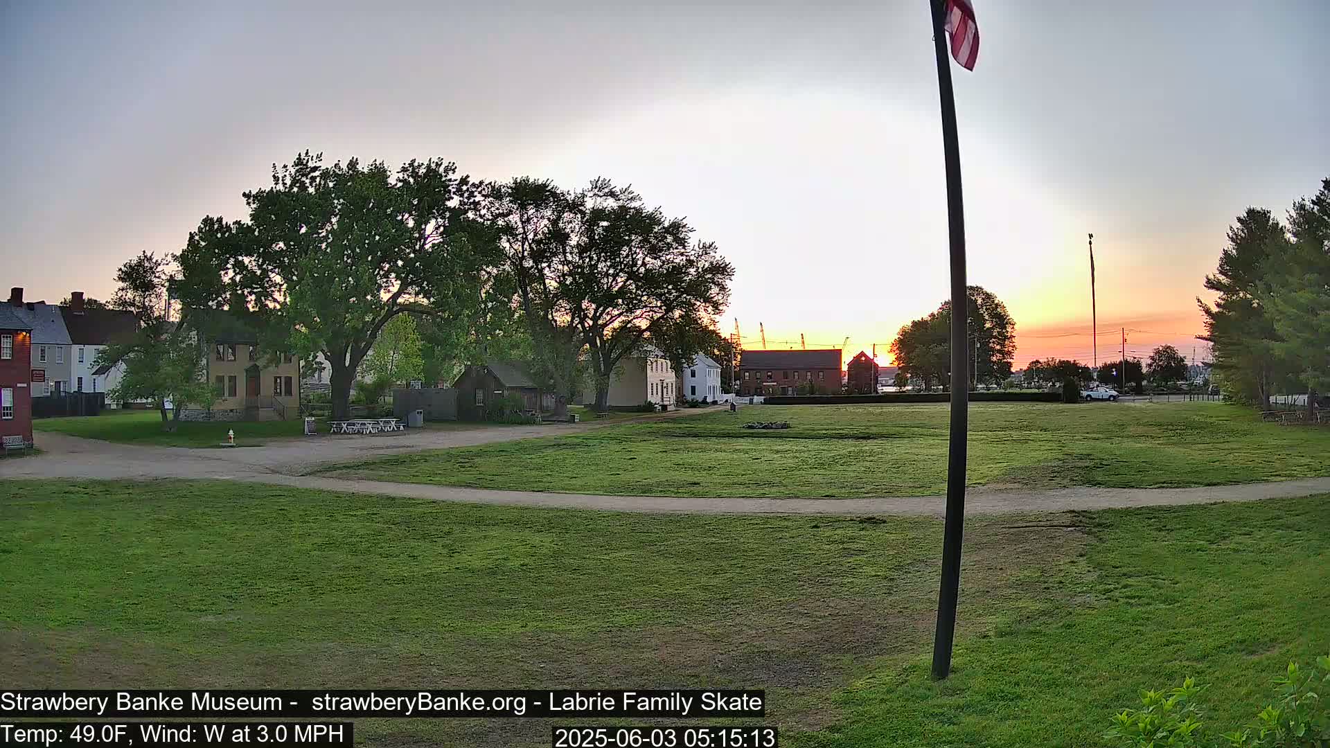 A grassy field with historical buildings and trees at sunset, under a clear sky with a light wind.