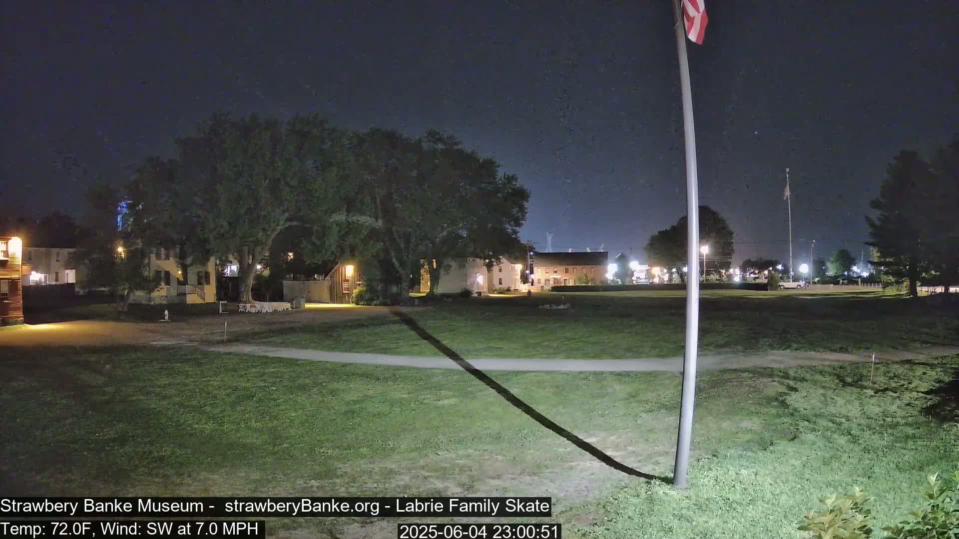 A nighttime view of a grassy area with several buildings in the background under a clear, starry sky.