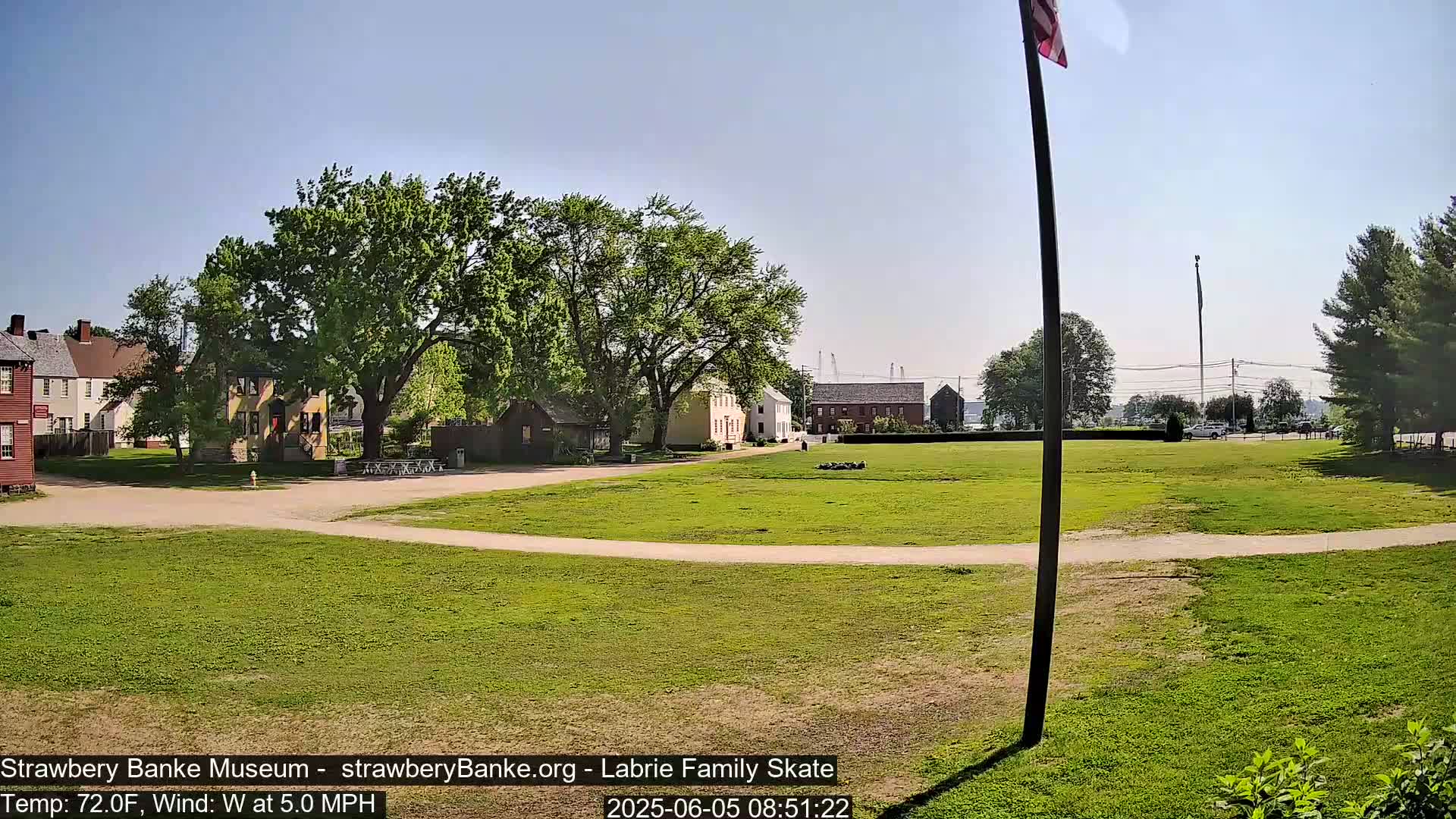 A grassy field with several historical-looking buildings and trees in the background on a sunny day with a 5 mph west wind and a temperature of 72°F.
