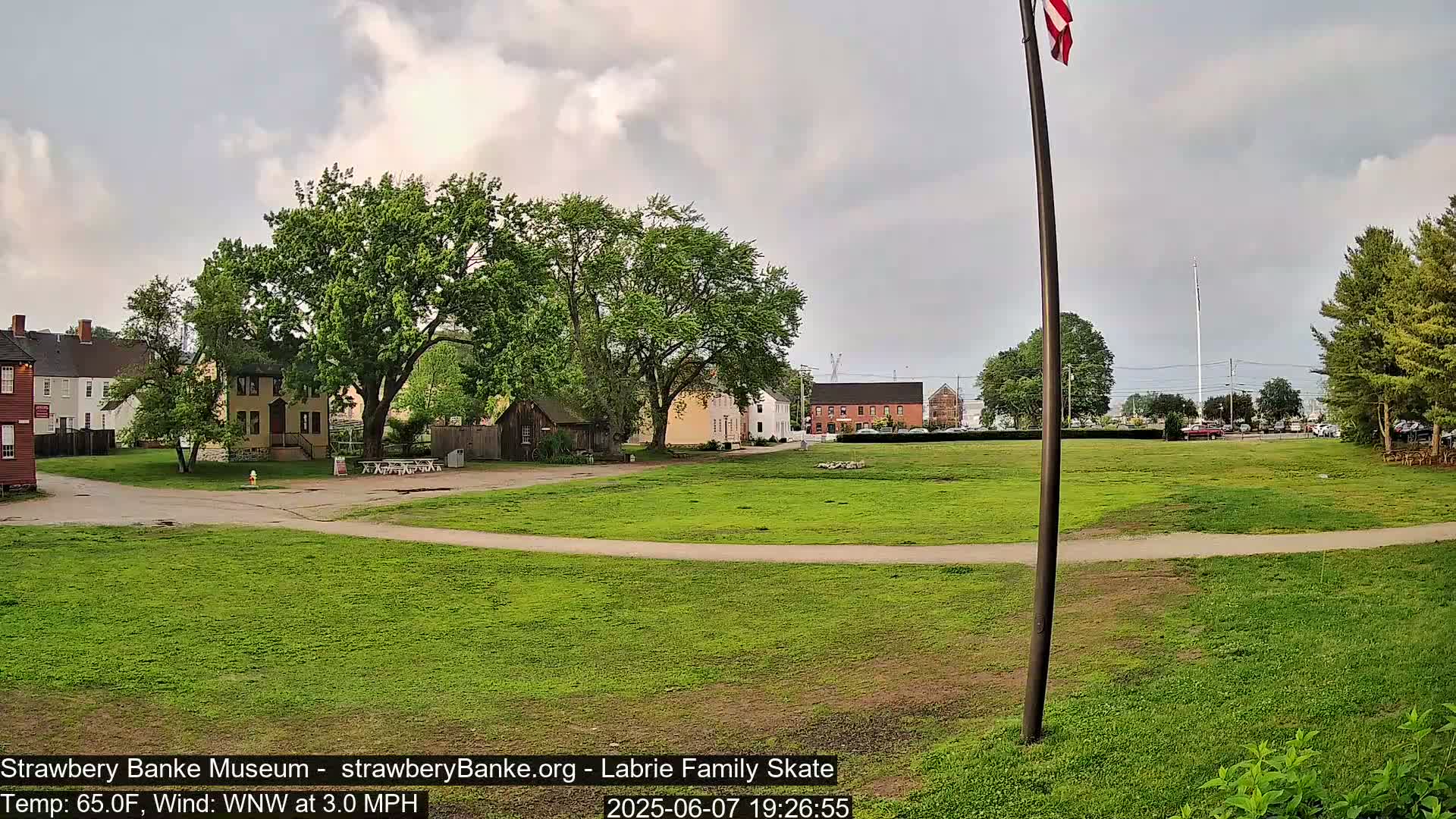 A grassy field with several historical-looking buildings and lush green trees under a partly cloudy sky.