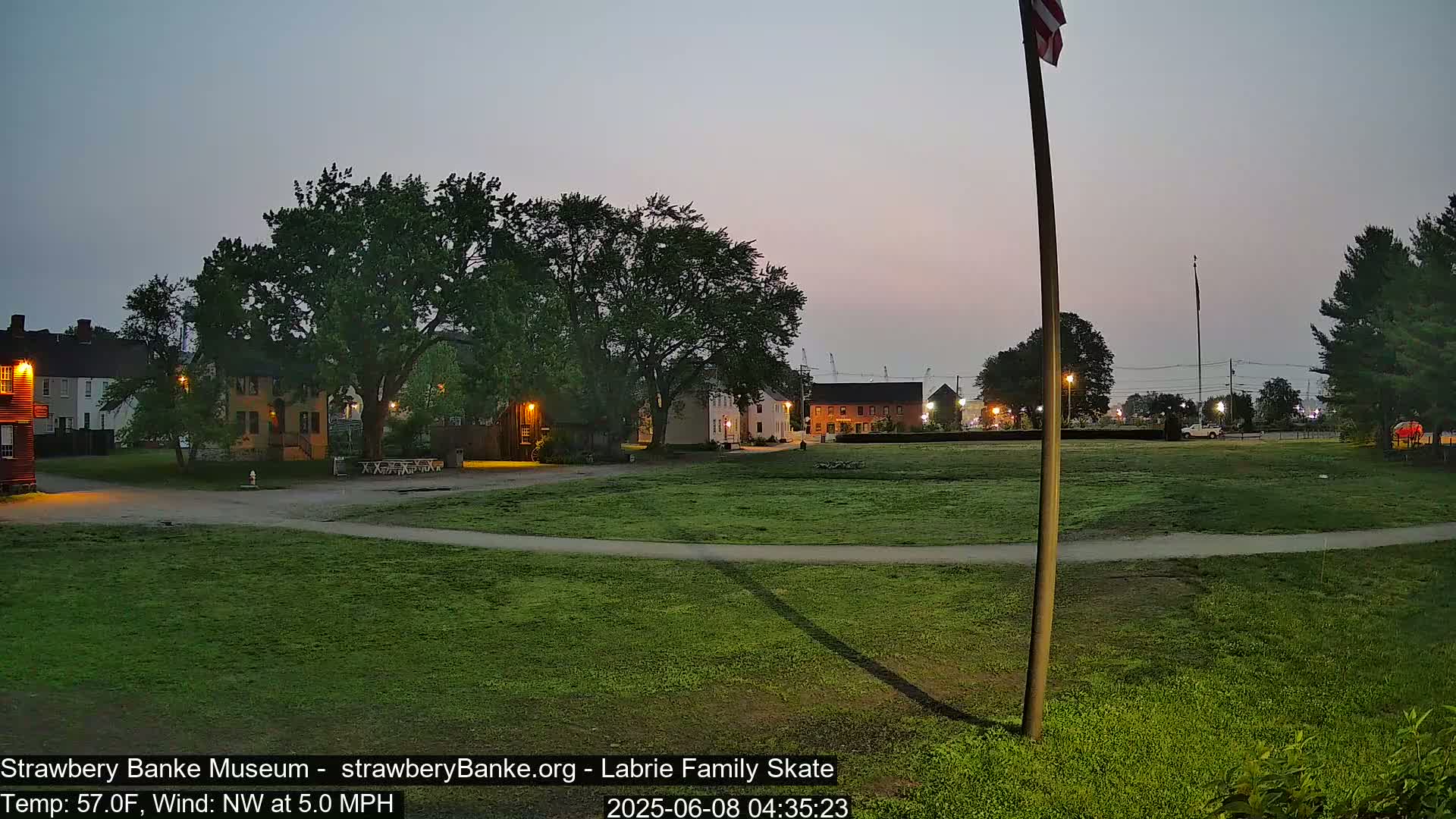 A grassy field at twilight, with several buildings and trees visible in the background, under a light grey sky.