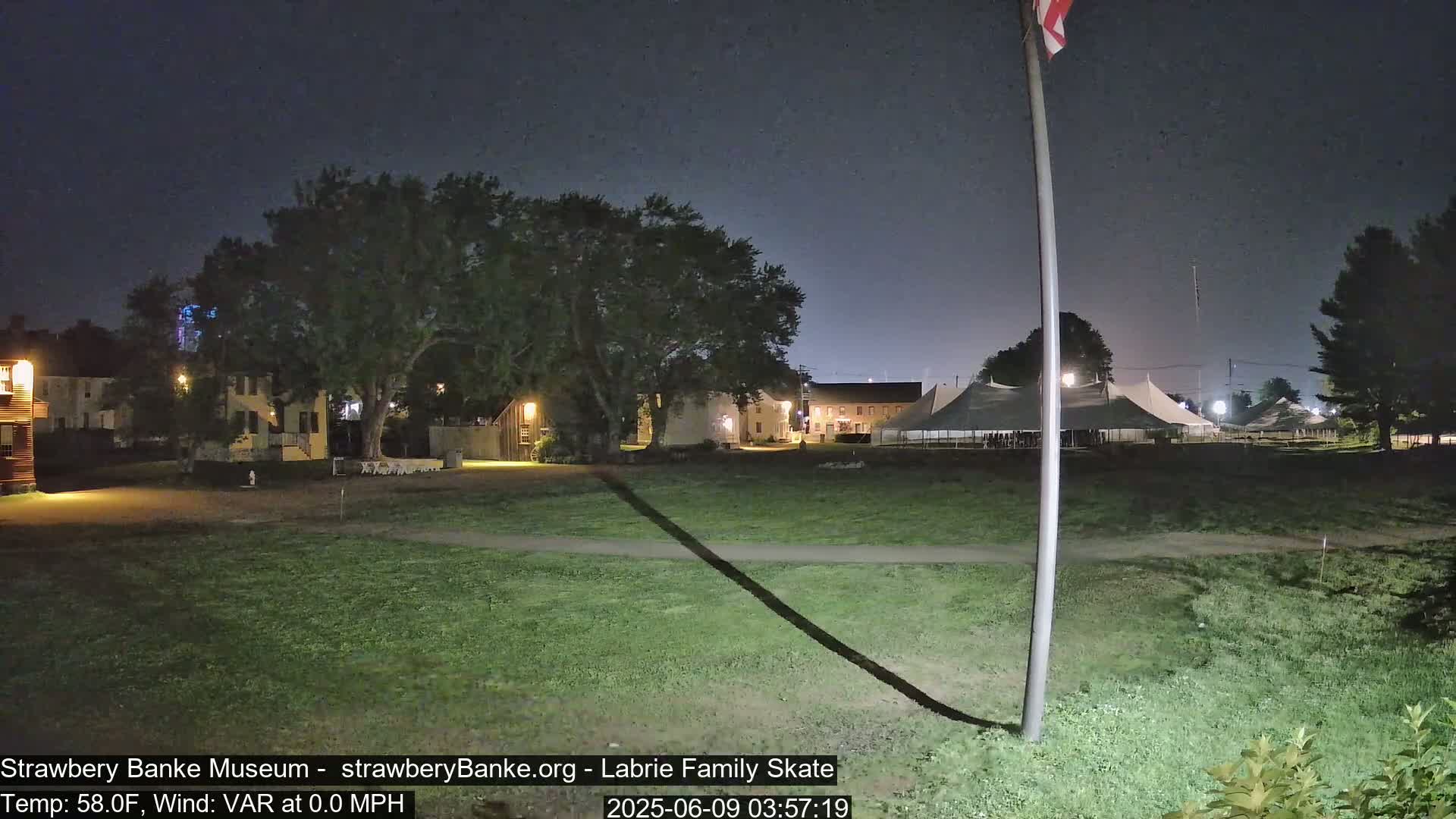 At night, under a dark sky, several buildings are illuminated, with a large tent and a flagpole in a grassy area.