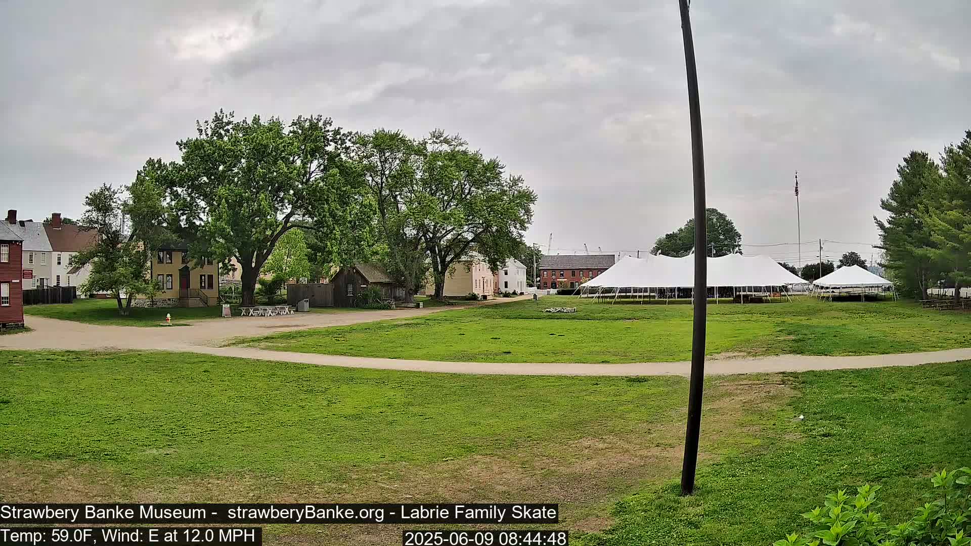 A grassy field with historical-looking buildings and large white tents is visible under an overcast sky, with an east wind blowing at 12 mph.
